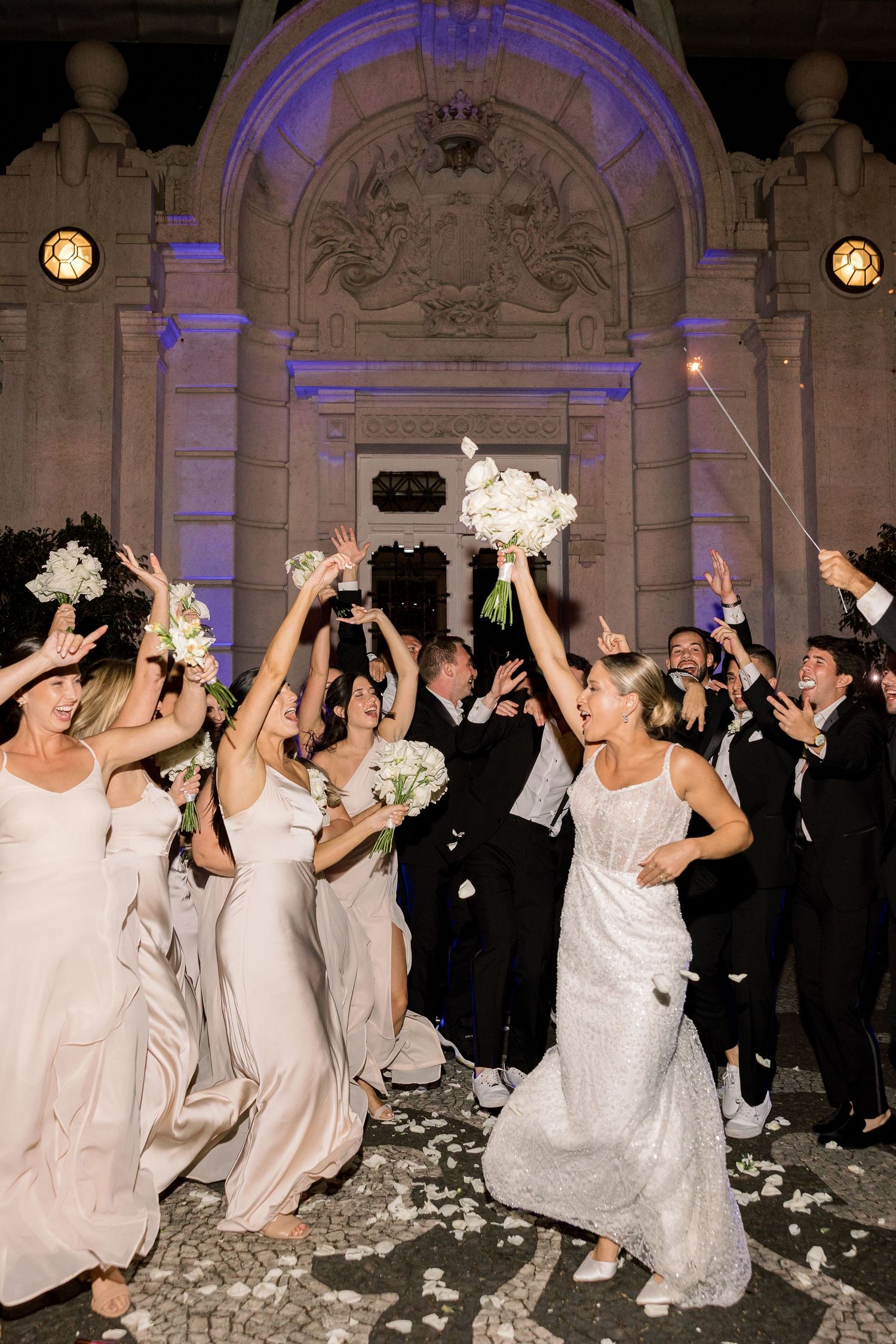 Wedding party in white dresses and black suits dancing energetically at an ornate venue.