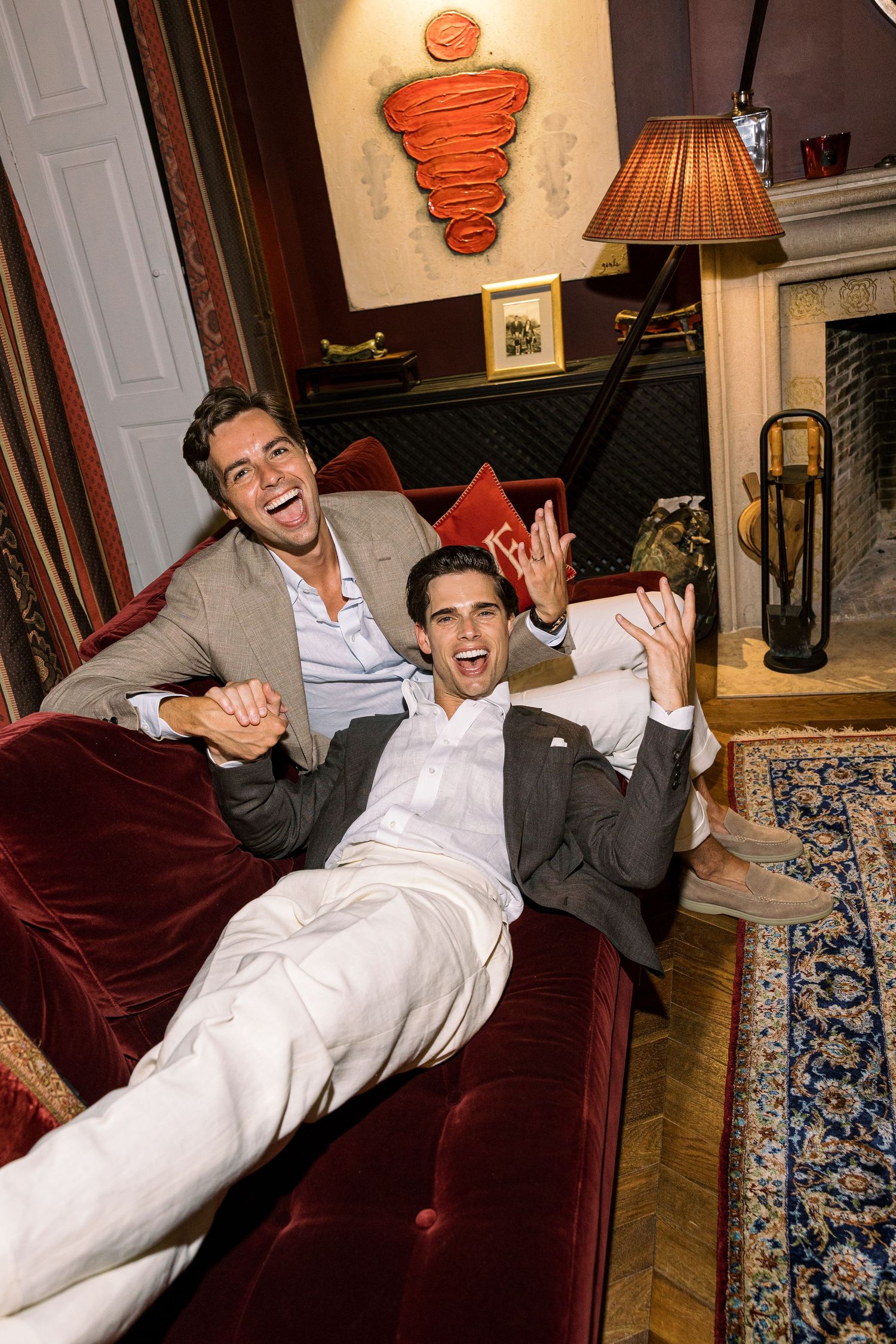 Two men in blazers laughing and celebrating on a red velvet sofa in an elegant, warmly lit living room.