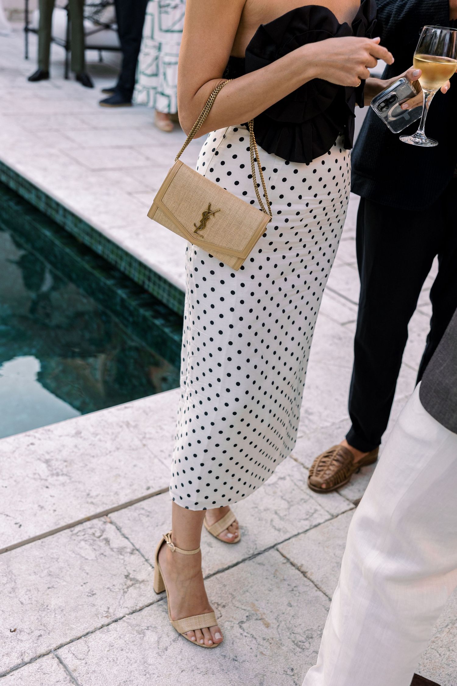 Woman in polka dot midi skirt holds champagne with YSL bag at elegant poolside event.