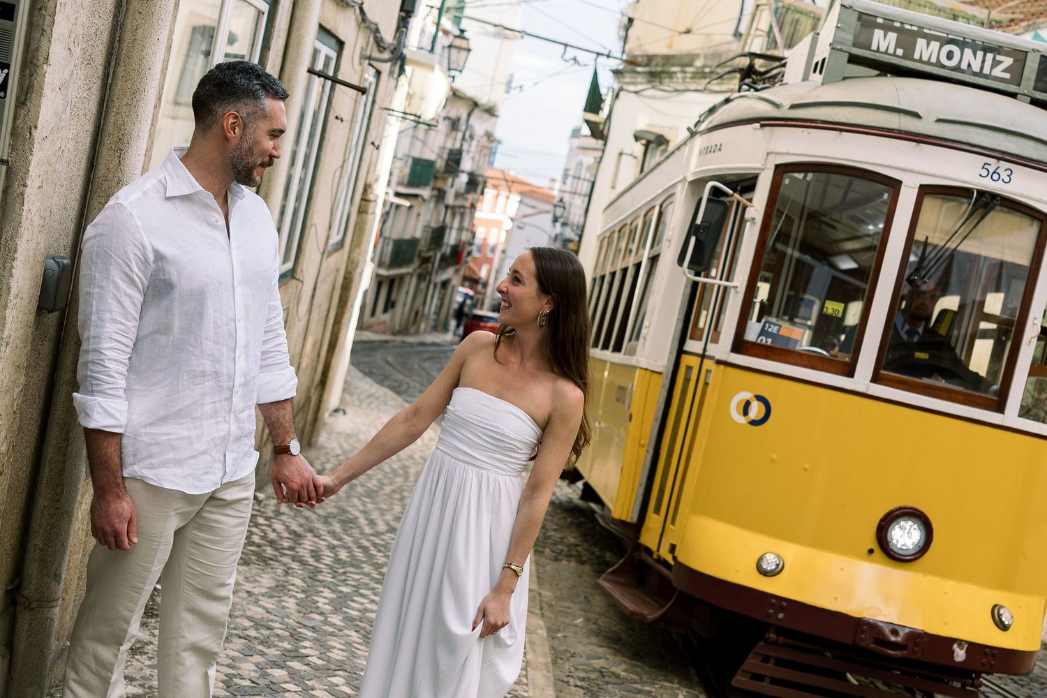 Couple holding hands near iconic yellow tram 28 on cobblestone street in Lisbon, Portugal.