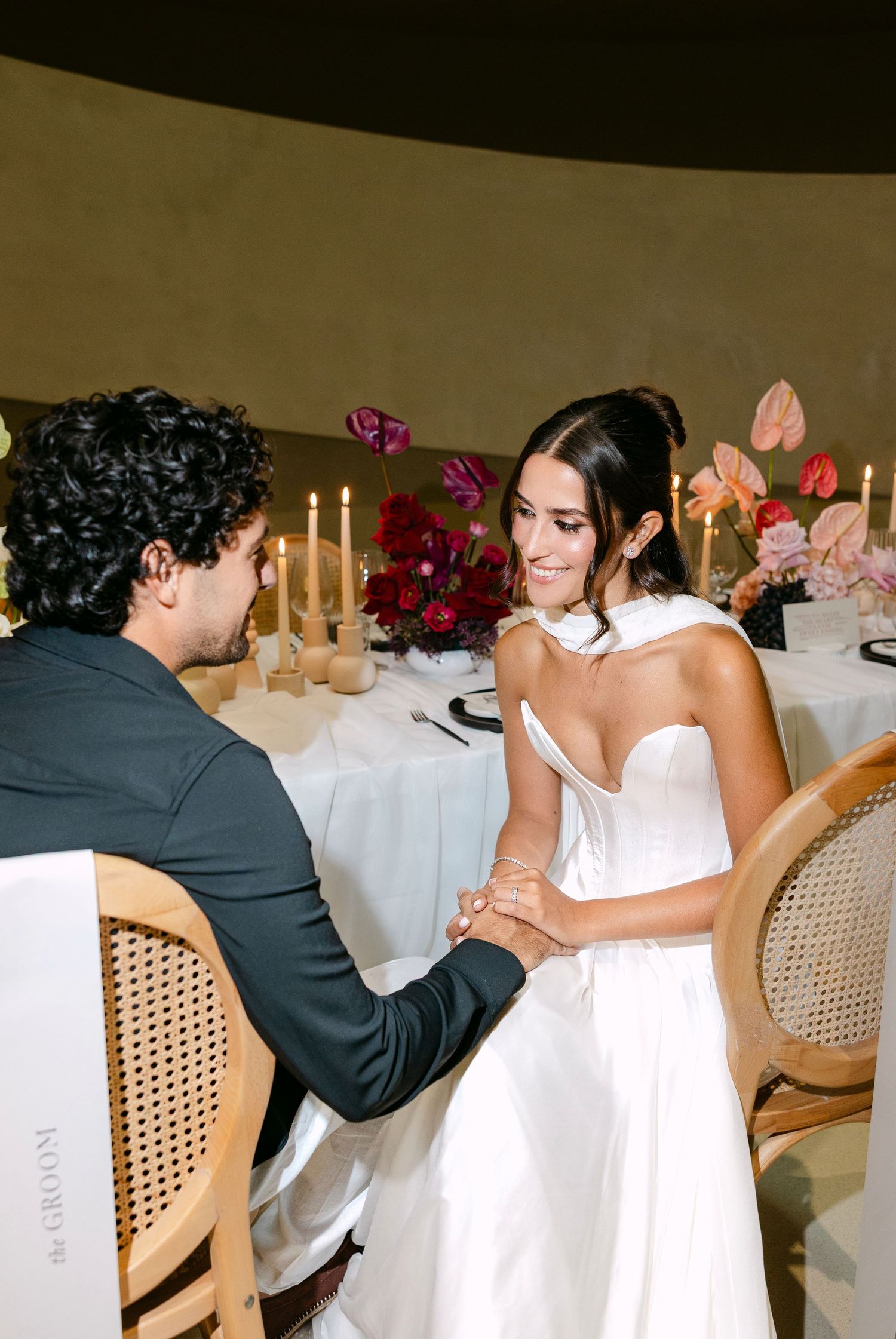 Bride in strapless white gown holds groom's hands at a candlelit wedding reception table with floral arrangements.