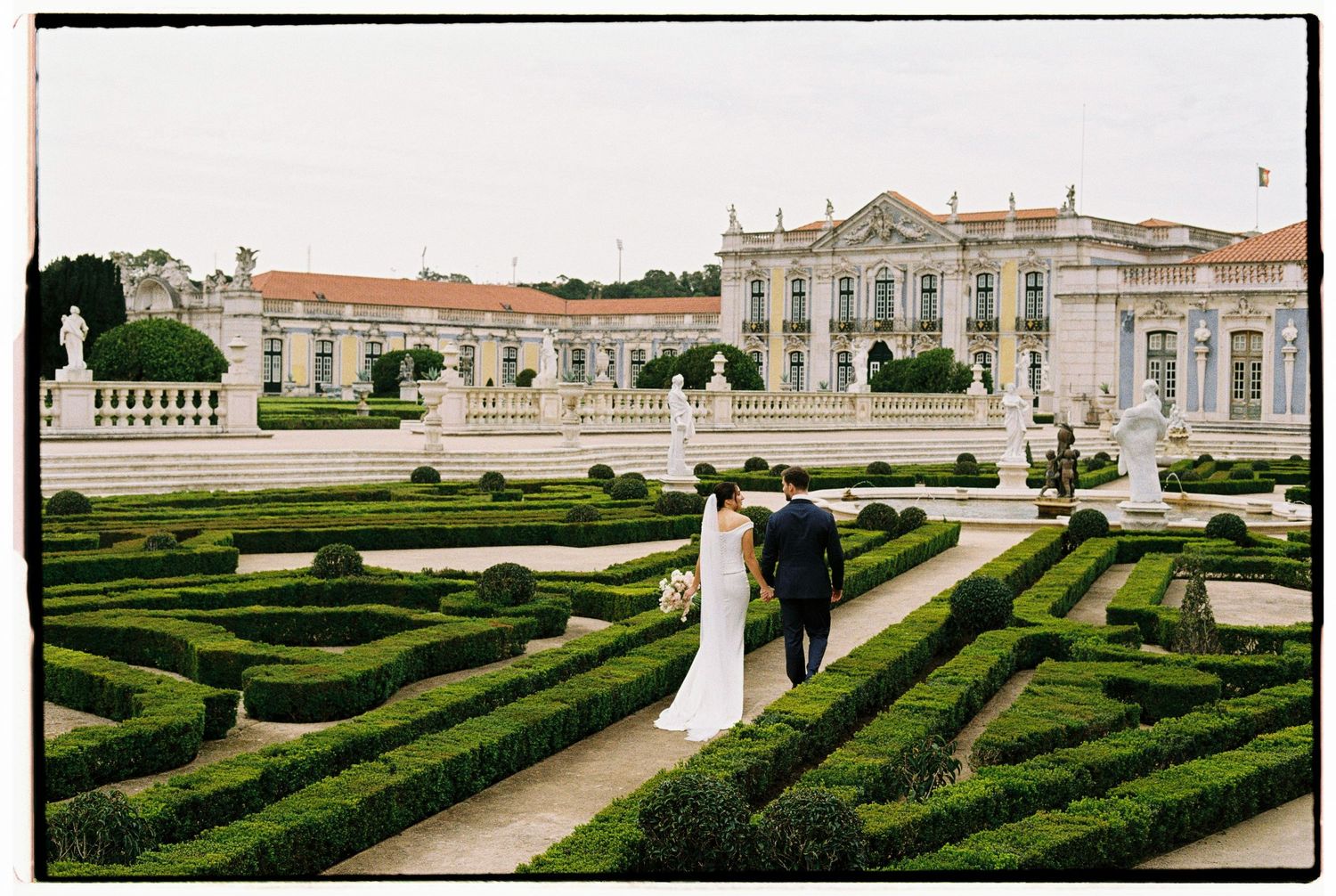 Bride and groom walk through manicured hedge gardens at an elegant Portuguese palace wedding venue.