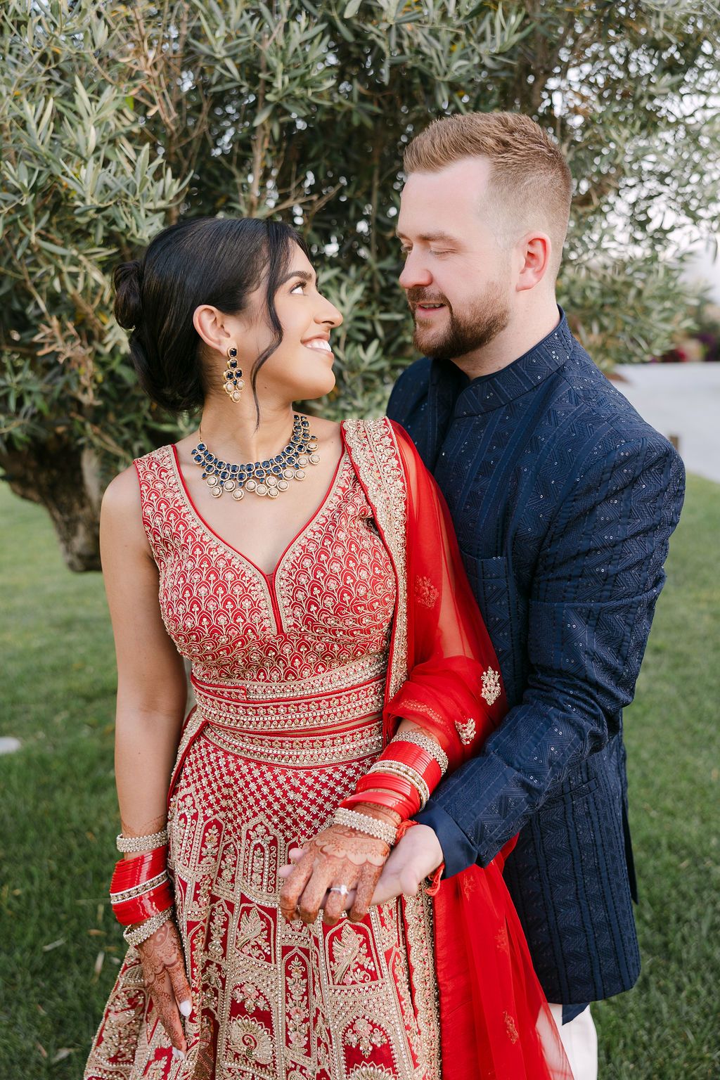 A couple in traditional wedding attire embraces outdoors, with the bride wearing an ornate red and gold lehenga.