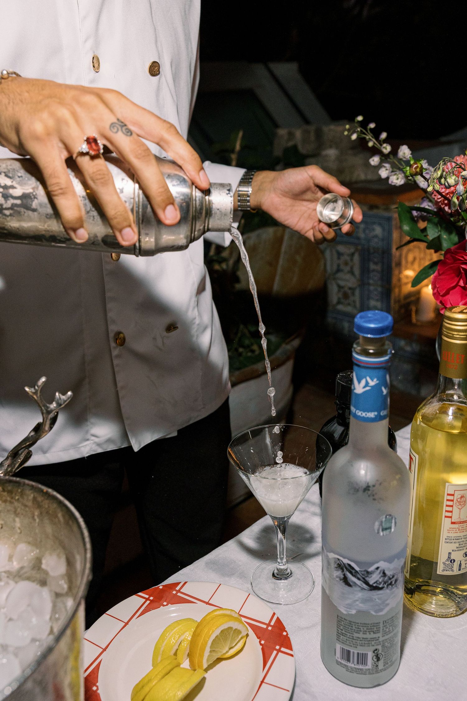 Bartender pouring cocktail from shaker into martini glass with Grey Goose vodka and lemon garnish on bar.