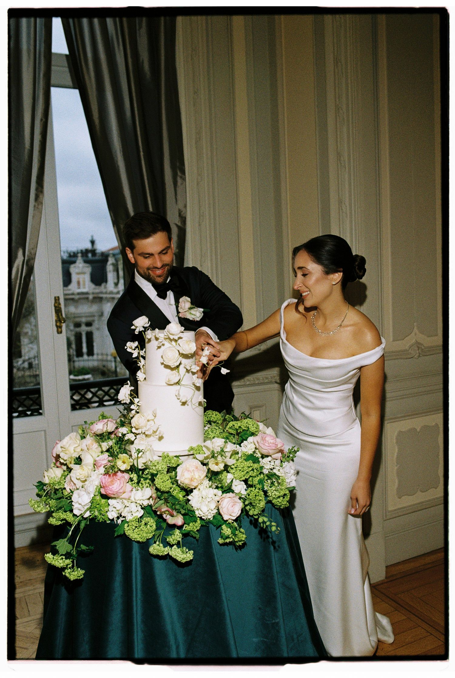 Bride and groom cutting white wedding cake adorned with flowers on green draped table in elegant venue.