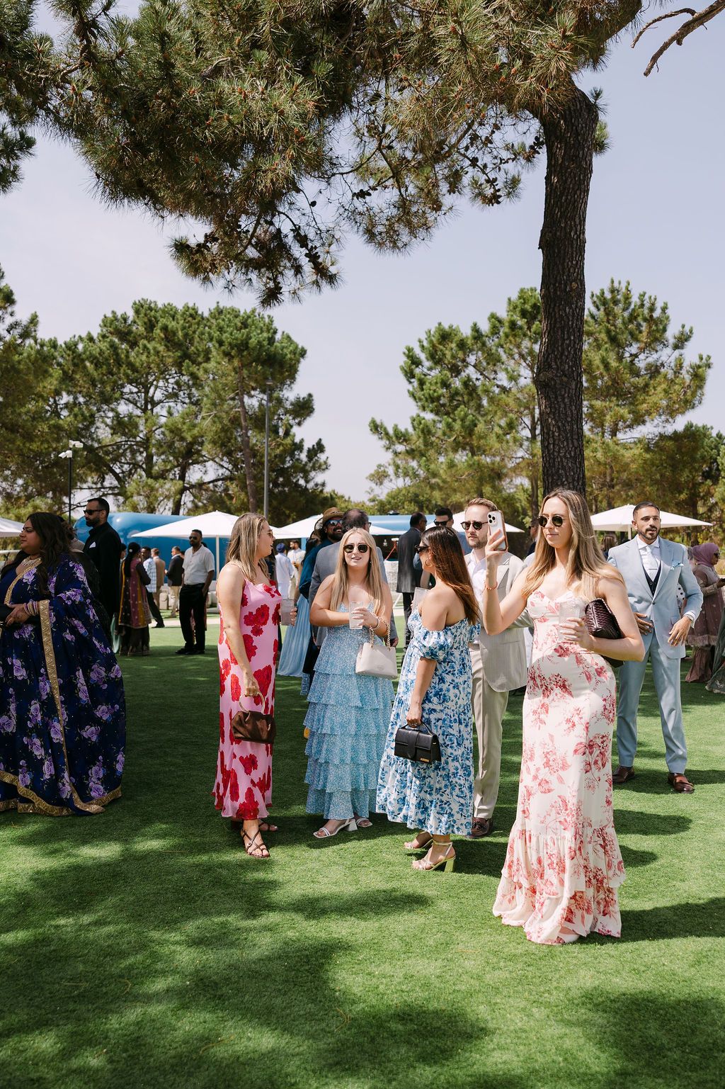 Wedding guests in colorful floral dresses gathered on a sunny lawn during an outdoor celebration.