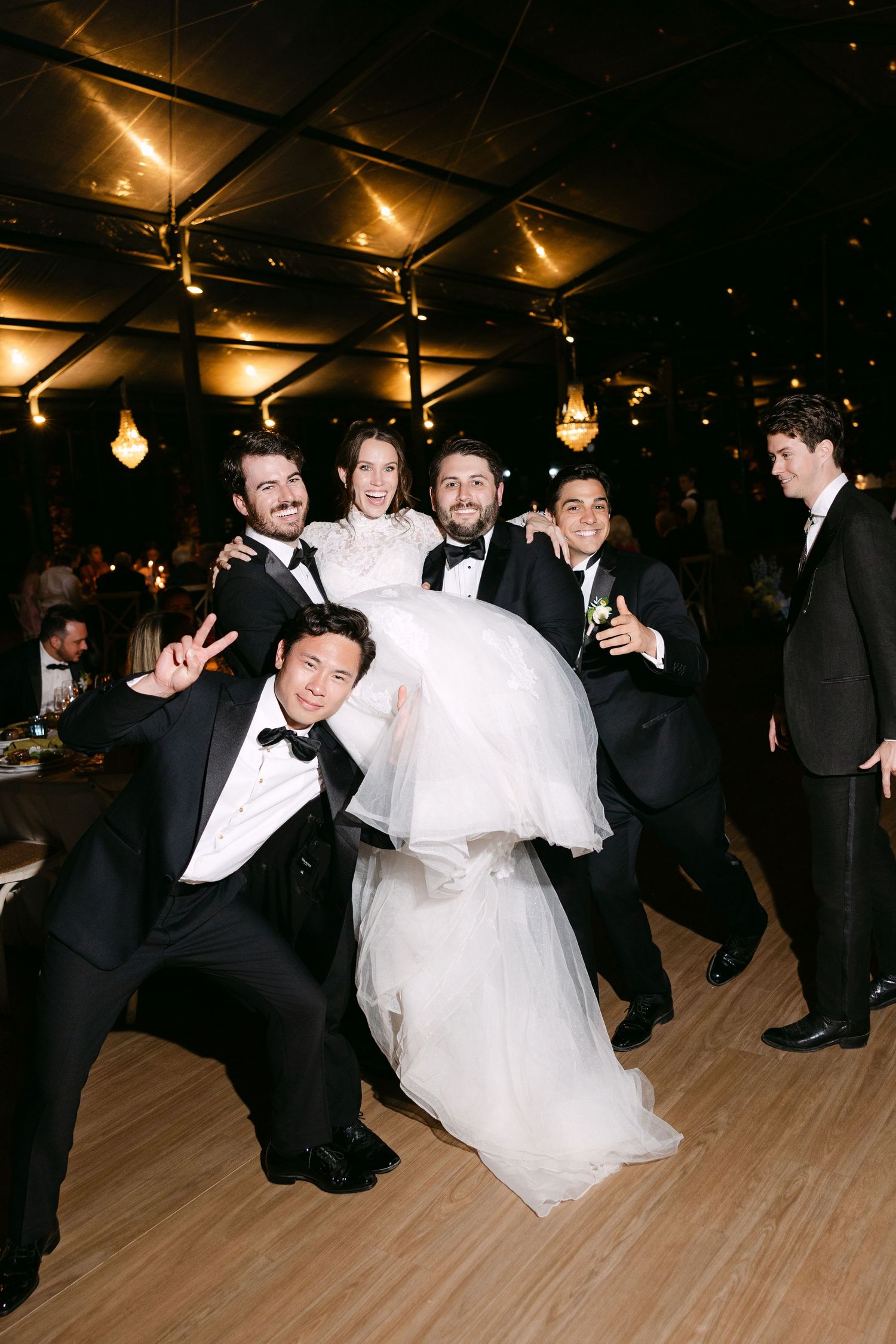 Wedding guests dance energetically on a wooden dance floor under warm lighting.