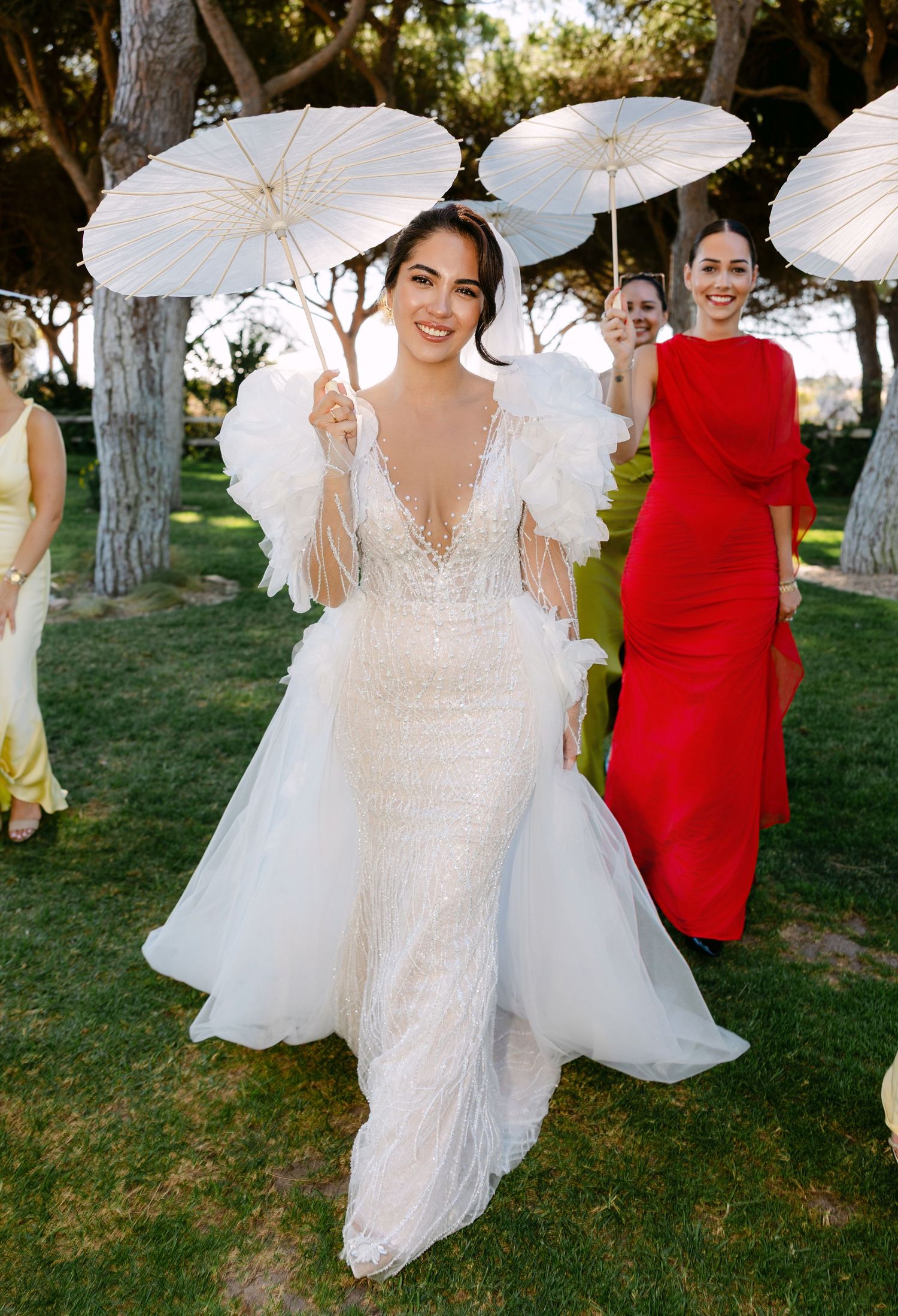 Wedding party members in white dresses and red gowns pose with parasols outdoors.