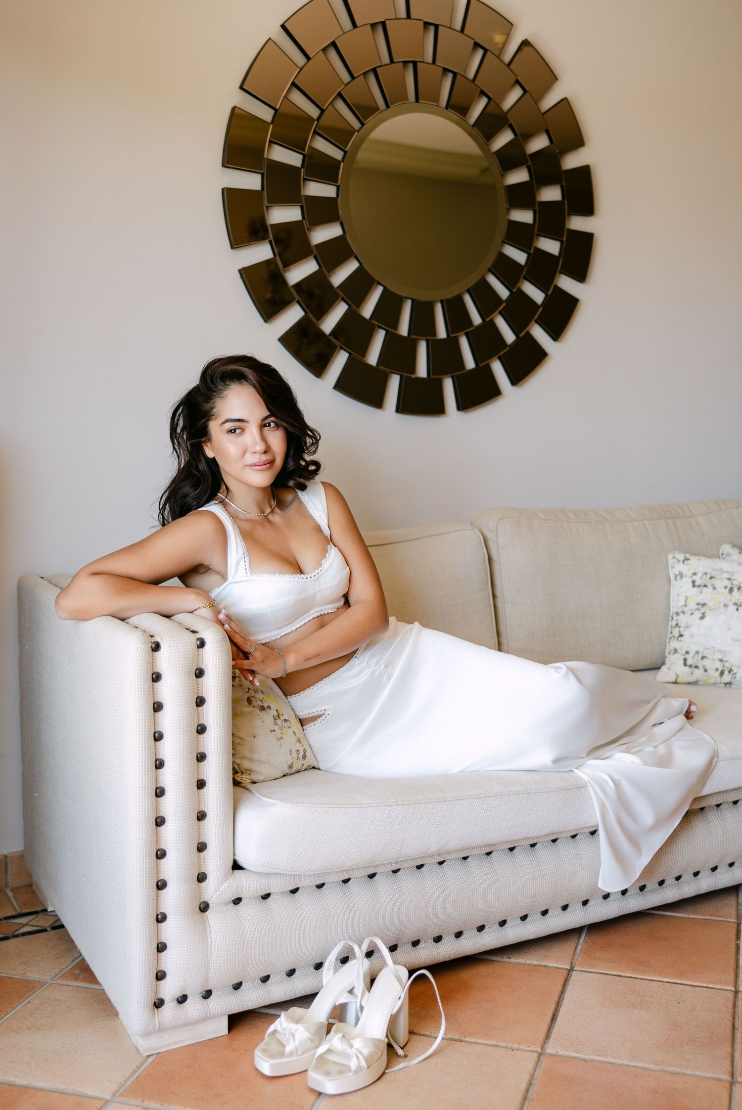 Wedding attire displayed on a white tufted chaise lounge beneath a sunburst mirror.