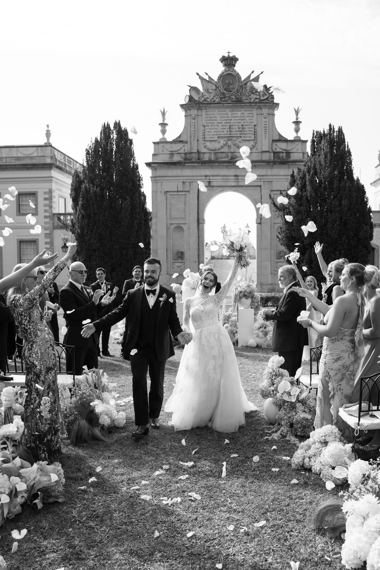 Wedding party celebrates with balloons in front of ornate white architectural gates.