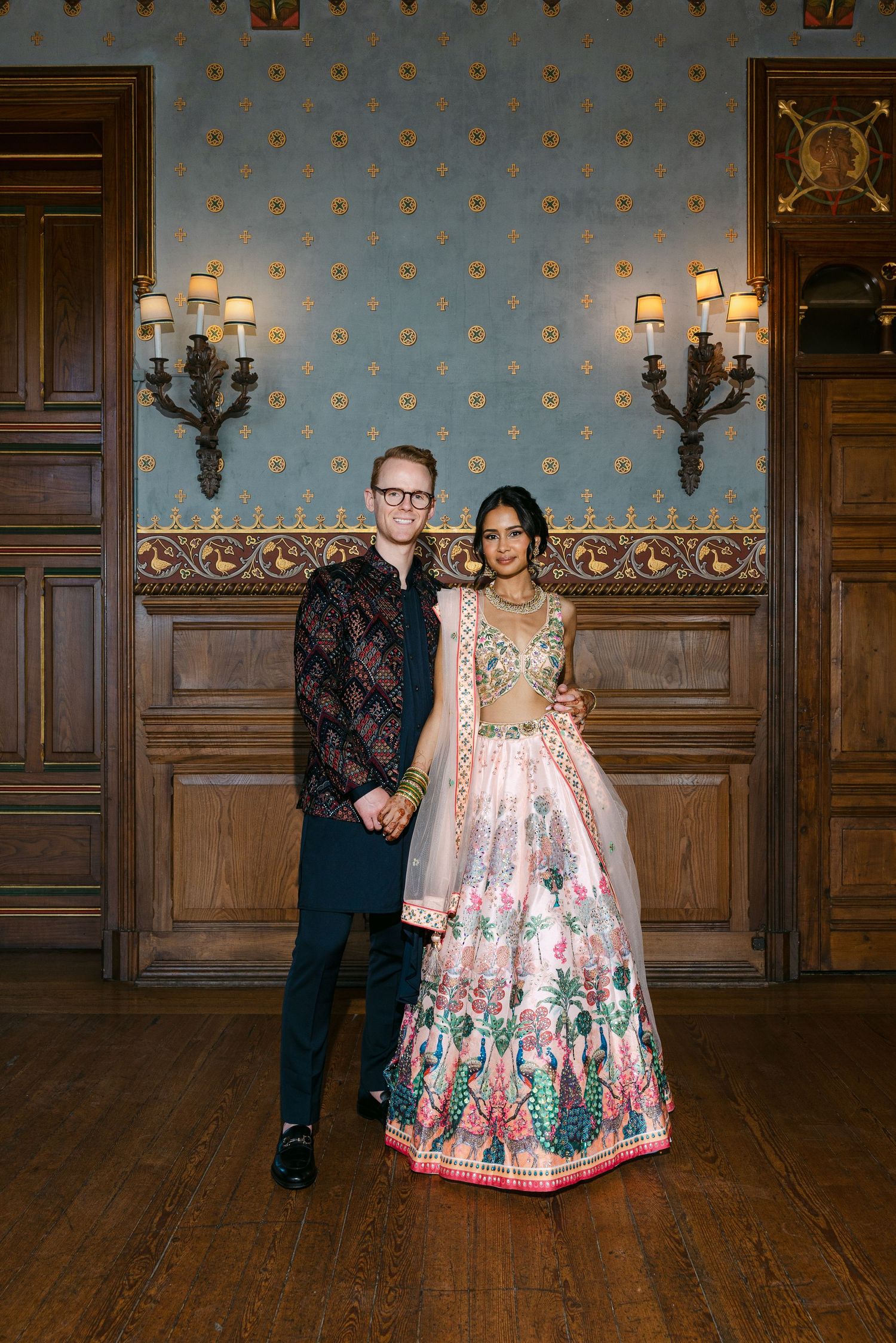 A couple poses in formal attire against an ornate blue wallpapered room with wall sconces.