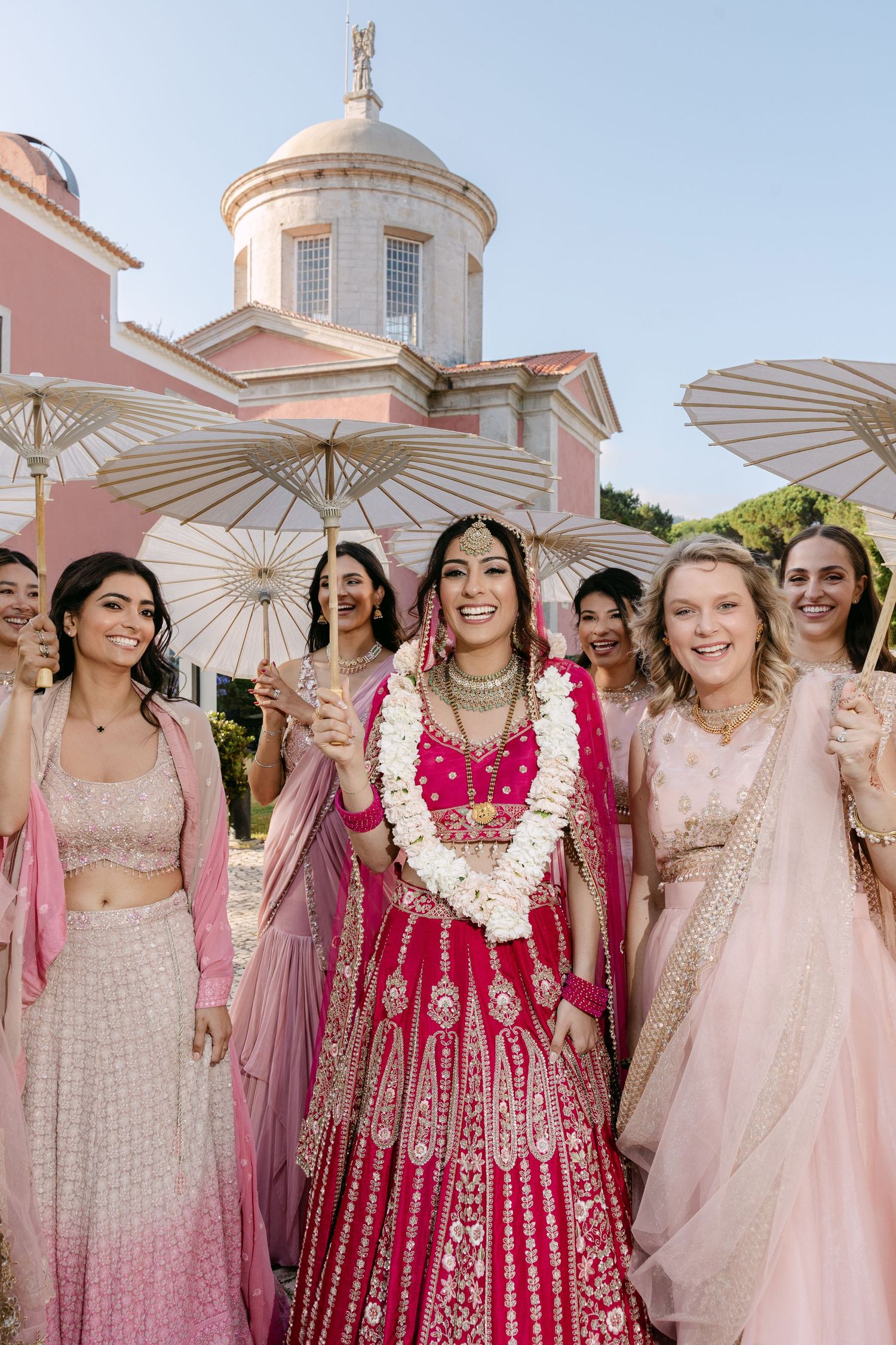 Wedding party with parasols posing in front of pink domed building in soft pink dresses.