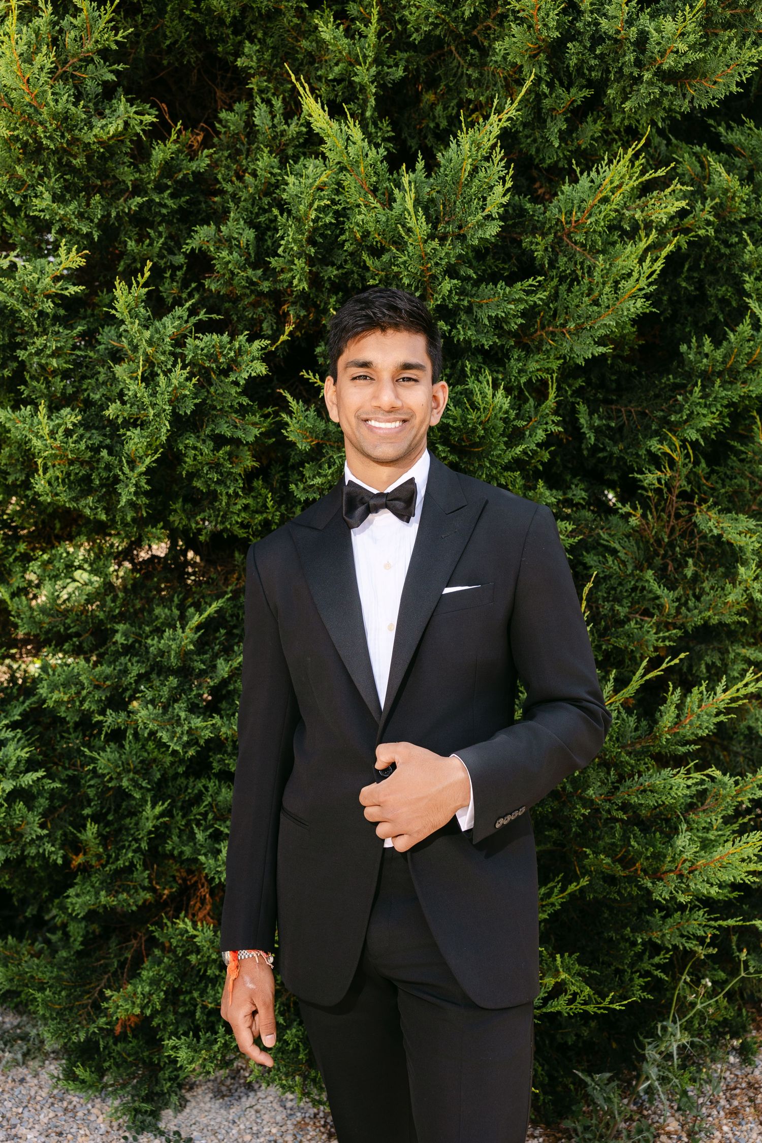 Formal portraits of groom in black tuxedo and bowtie against green hedge backdrop.