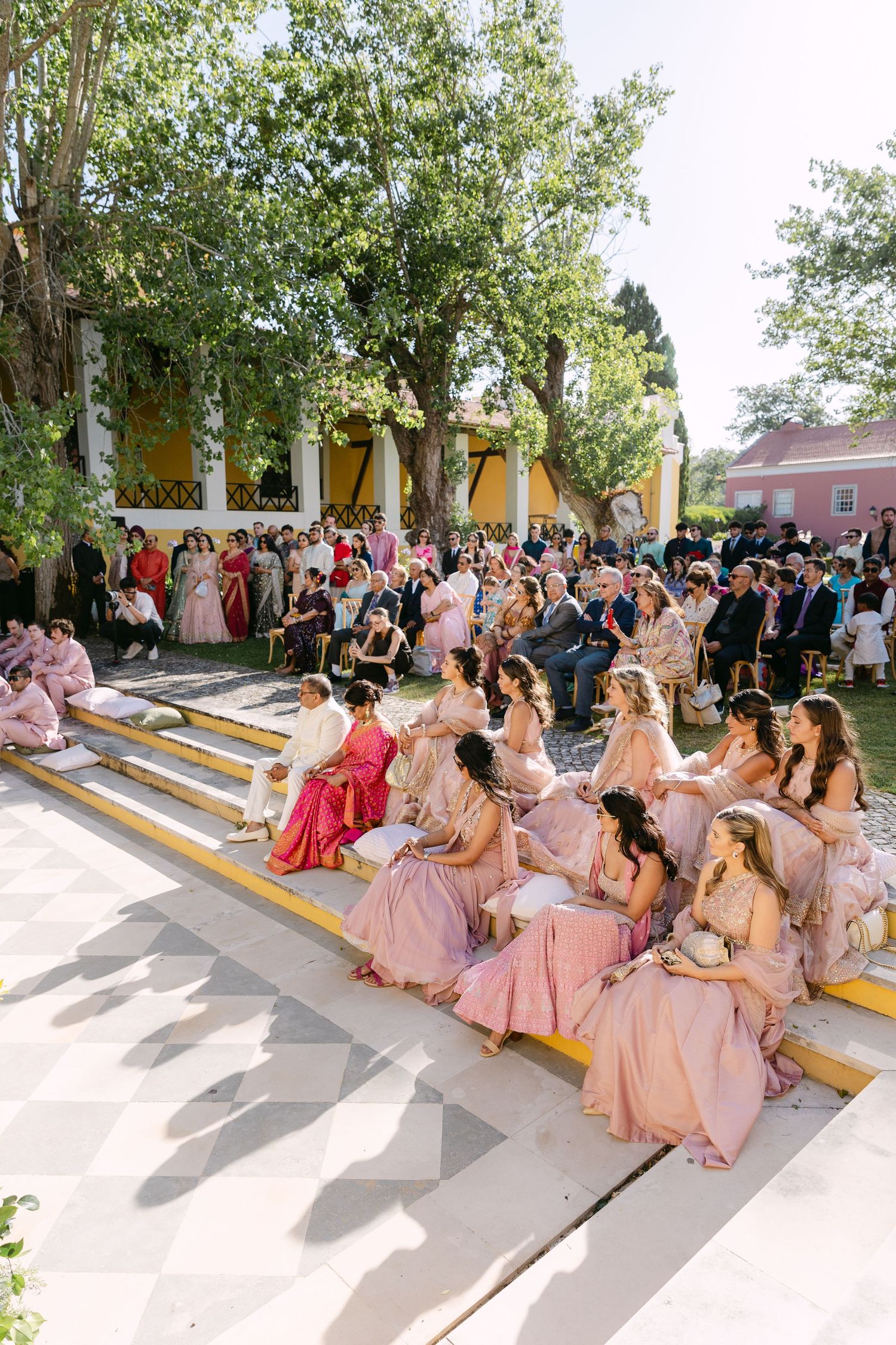 Wedding party members in pink dresses seated on outdoor steps for a group photo.