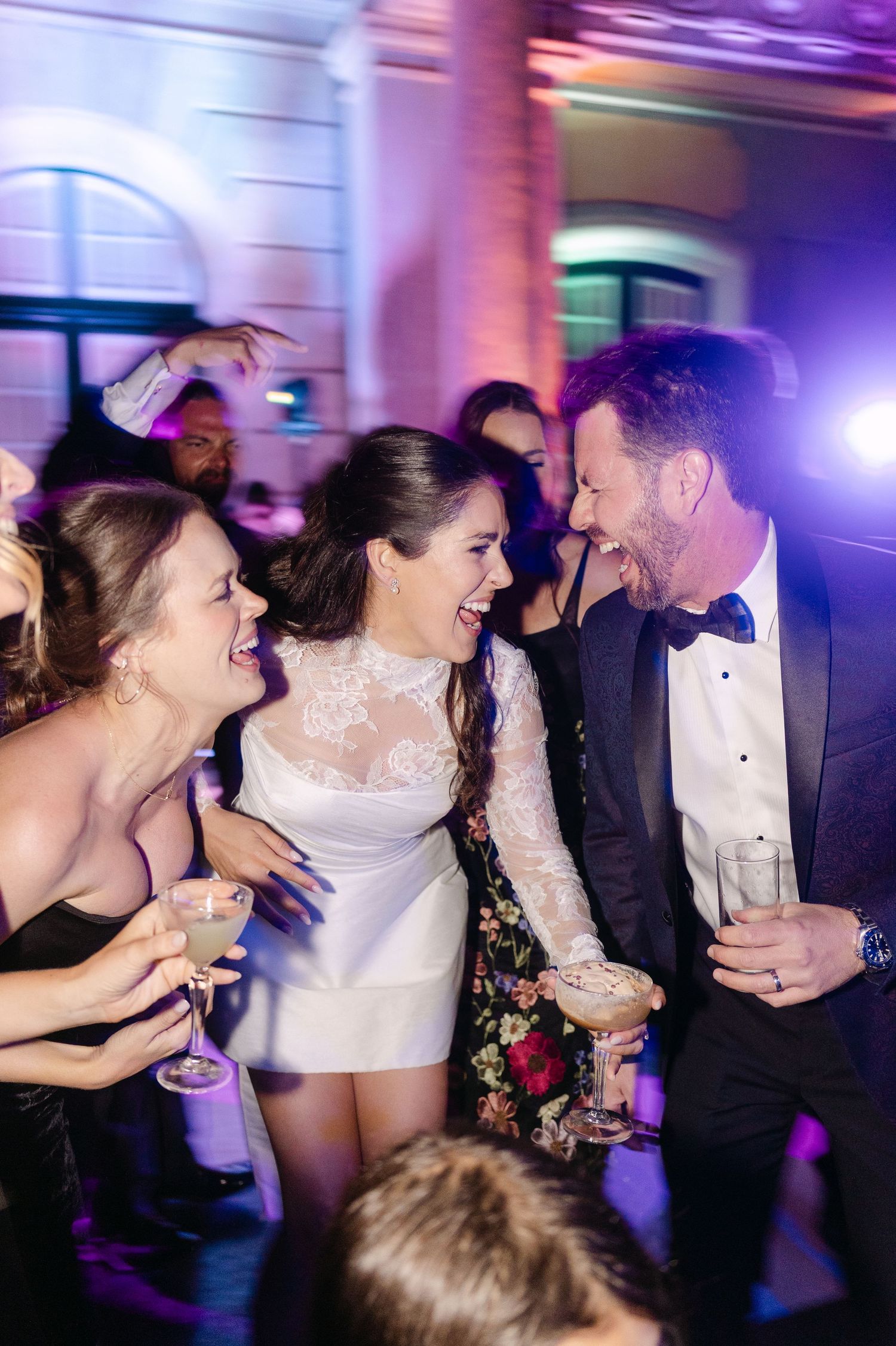 Bride in white mini dress laughing with guests on a colorfully lit wedding reception dance floor.