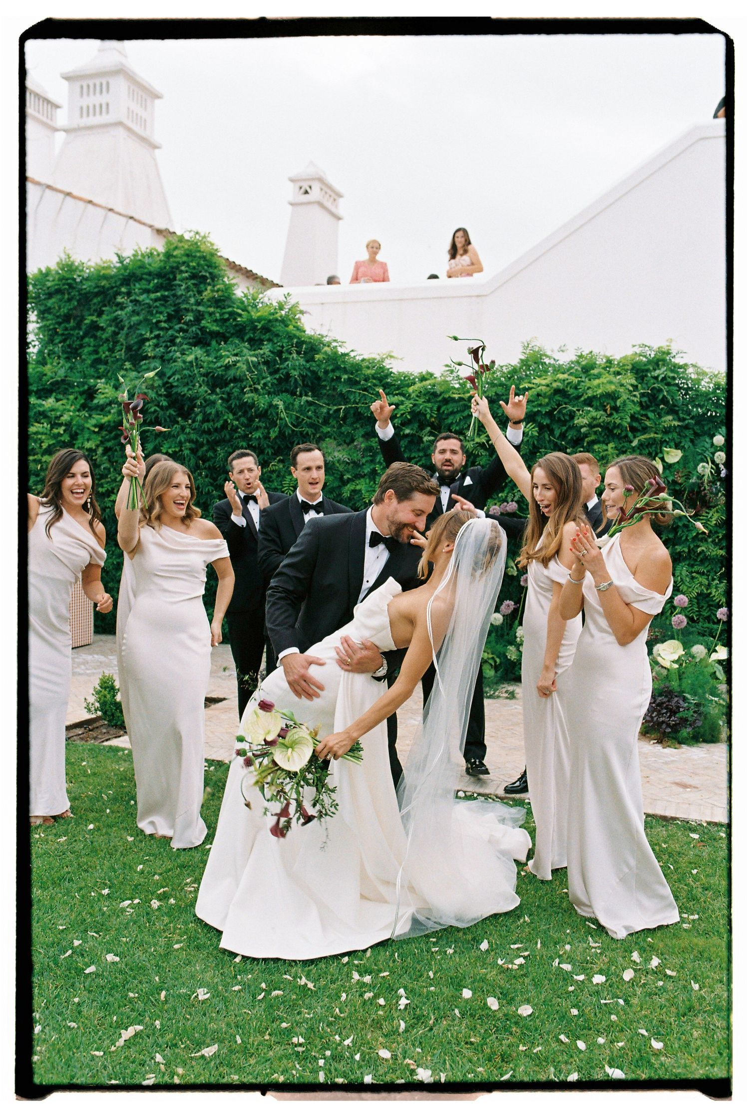 Bride and groom share a dip kiss surrounded by cheering wedding party in white gowns and black tuxedos on lush garden lawn.
