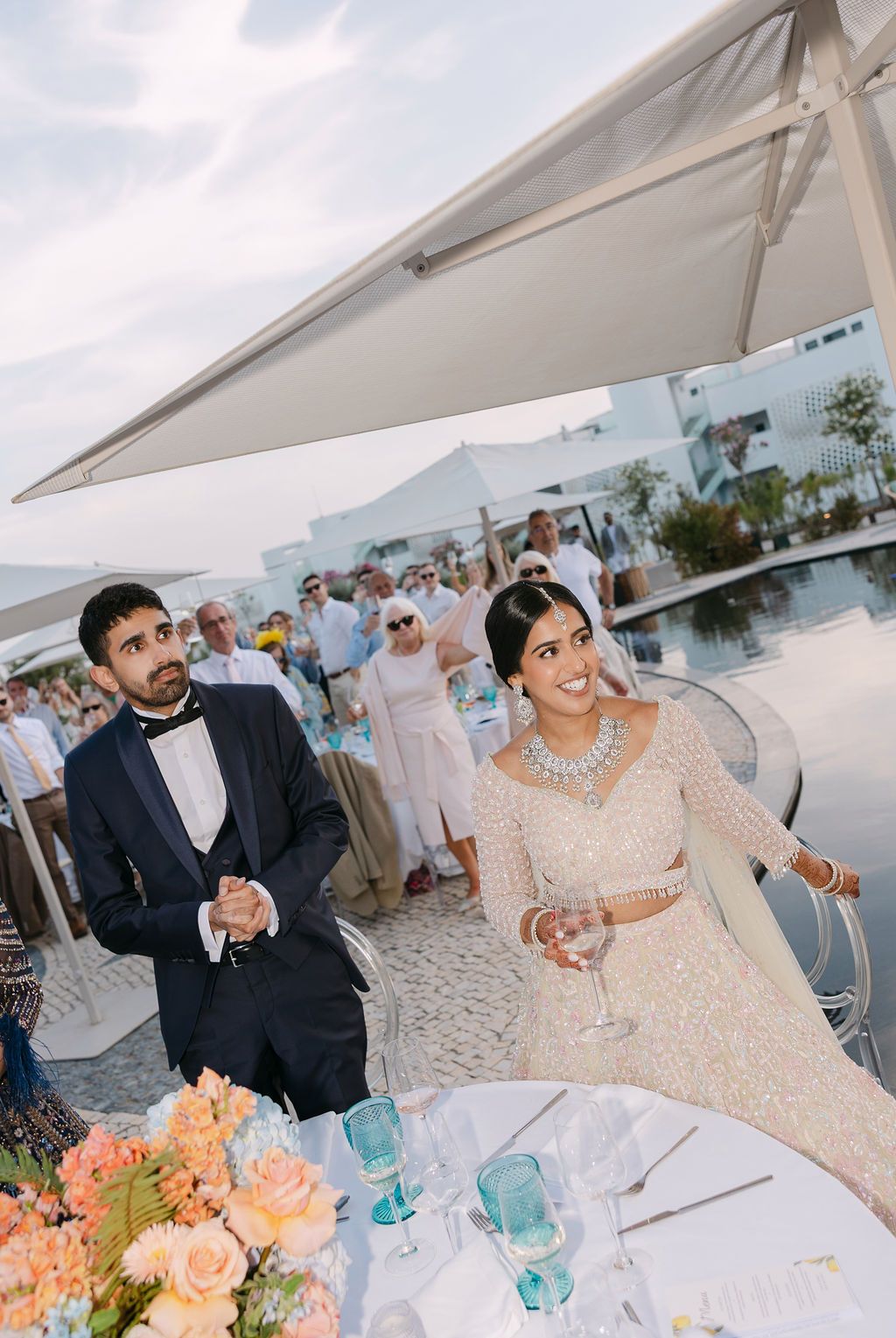 Wedding reception celebration under white canopy with floral centerpieces and coastal backdrop.