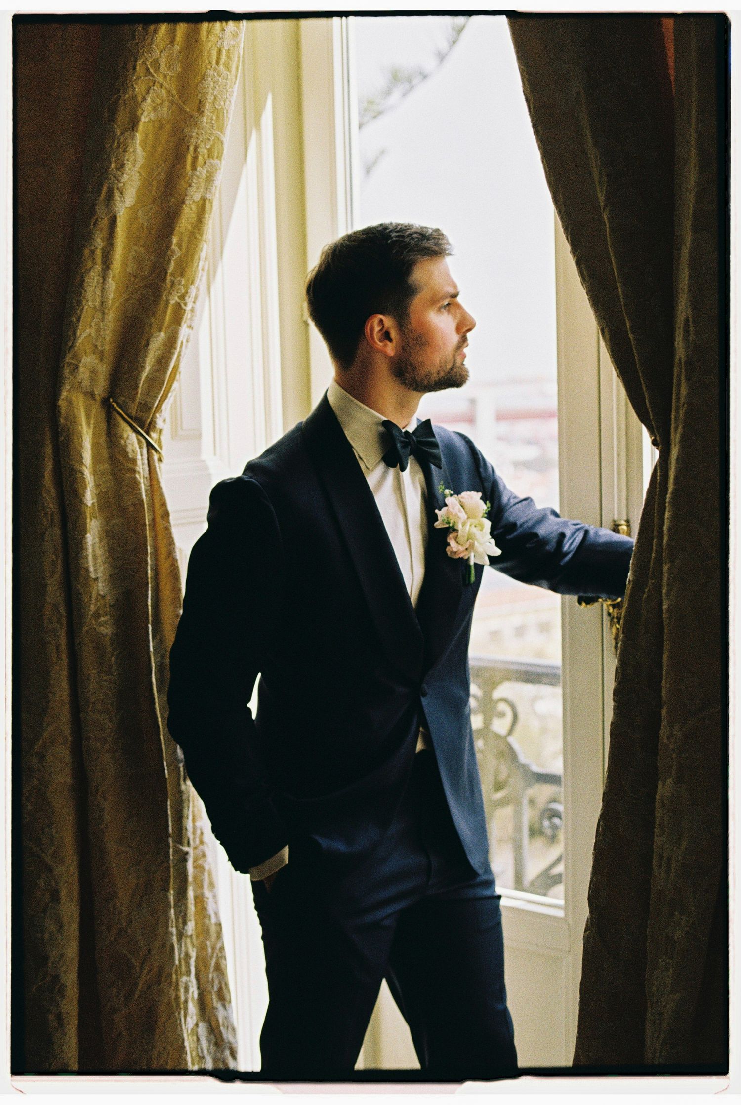 Groom in navy tuxedo with boutonniere gazes out window between elegant curtains on wedding day.