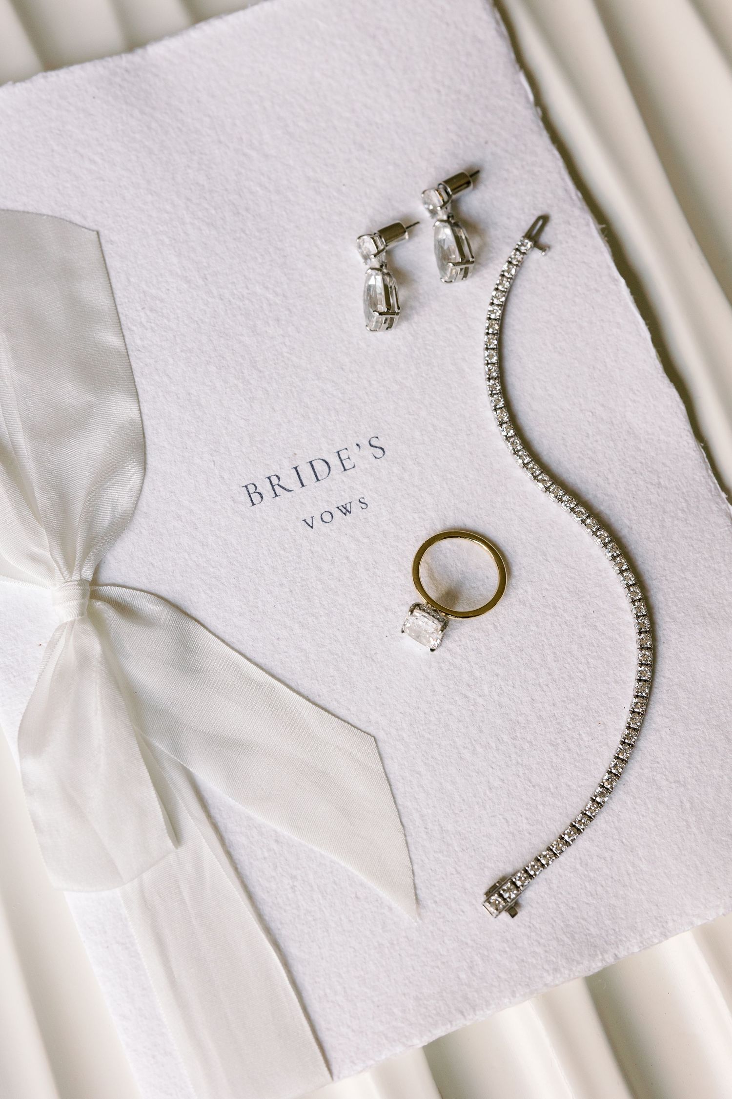 Wedding accessories including diamond jewelry and vows book arranged on white textured paper.