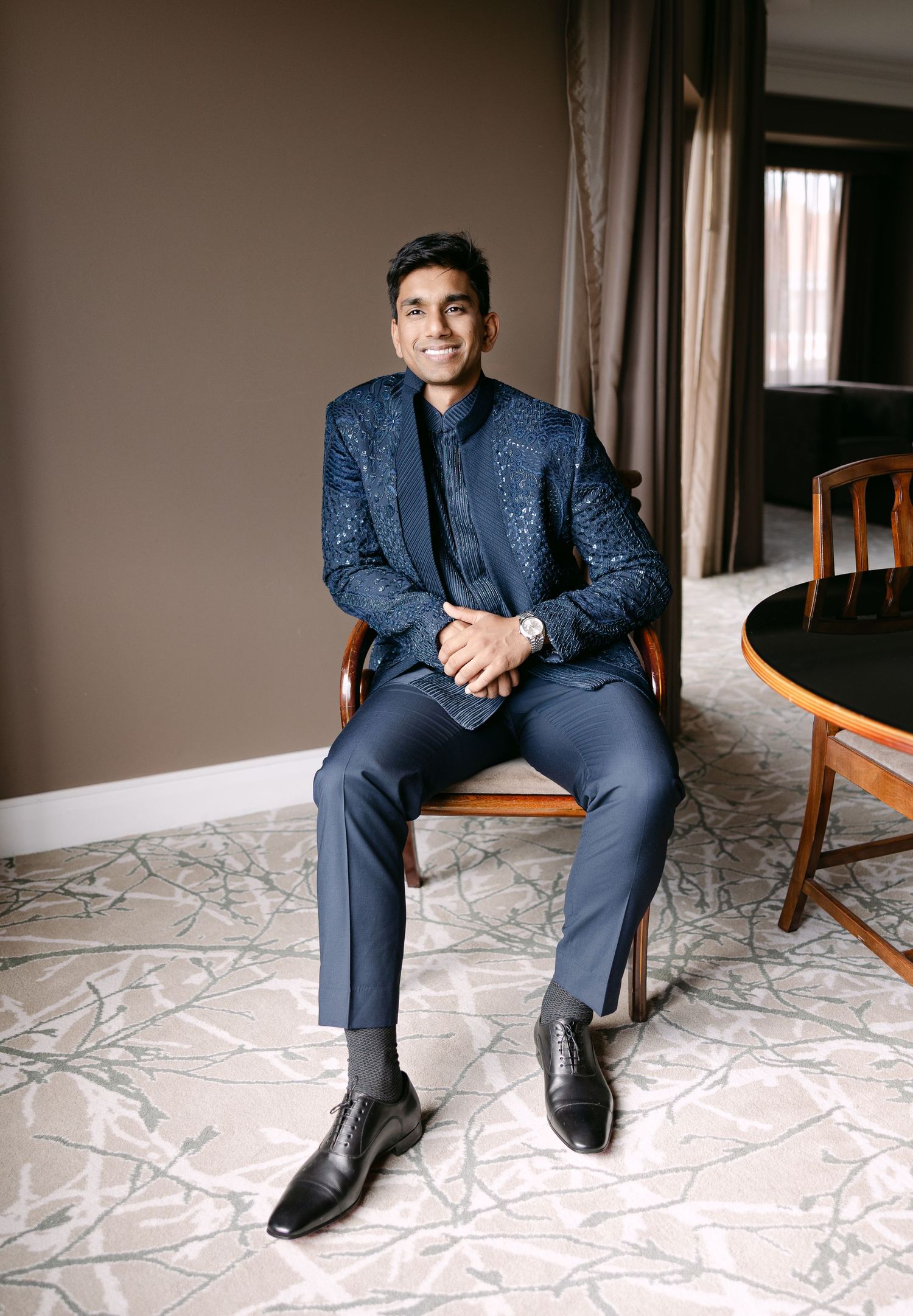 Person in navy patterned suit sits elegantly on chair against neutral background.