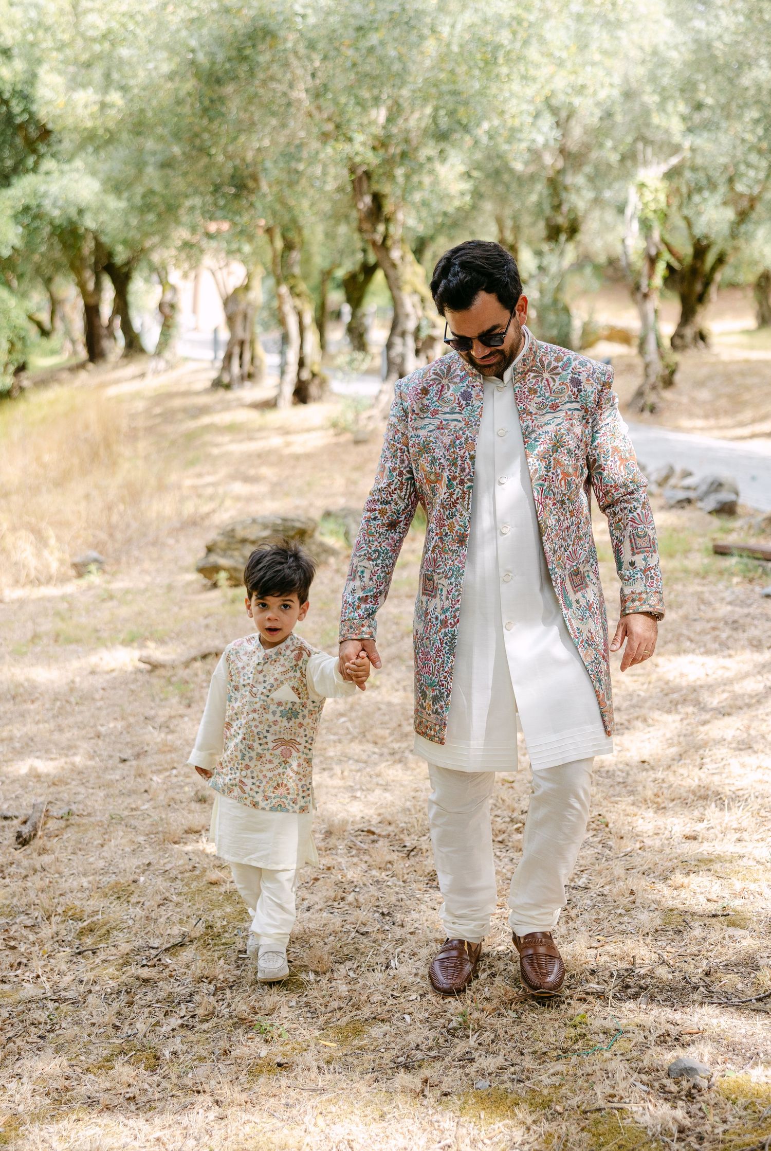 Father and child walk together in an olive grove wearing matching floral print jackets and white outfits.