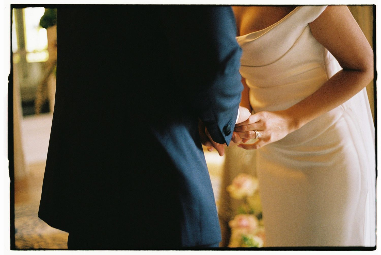 Bride and groom holding hands at wedding ceremony, bride in elegant white gown, groom in navy suit.