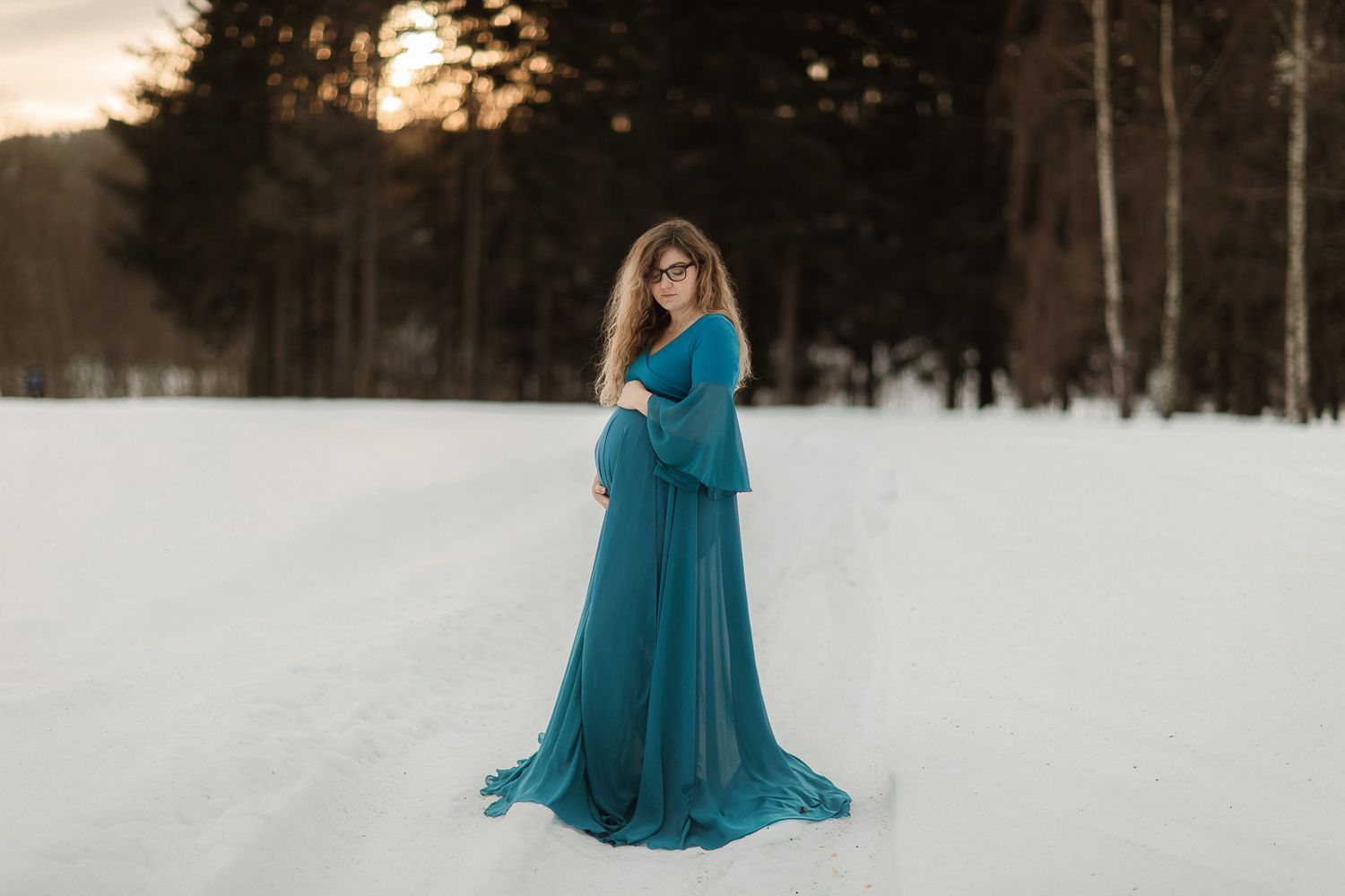 En gravid kvinna klädd i blå bohemisk klänning, som står på snön, med skog och en solnedgång i bakgrunden.