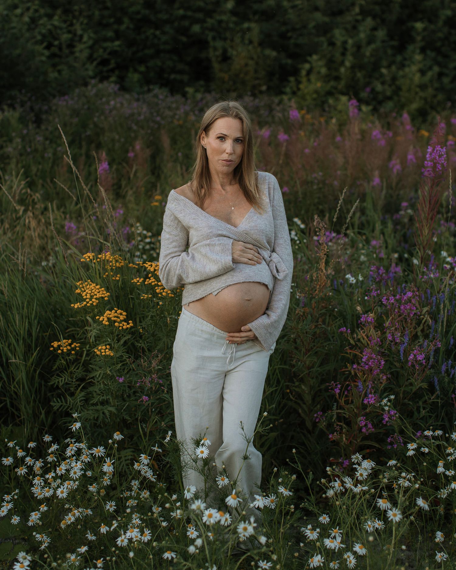 Gravid kvinna på en blomsterfylld äng håller om sin mage. Hon är klädd i en grå tröja och vita byxor och har ett lugnt, harmoniskt uttryck.