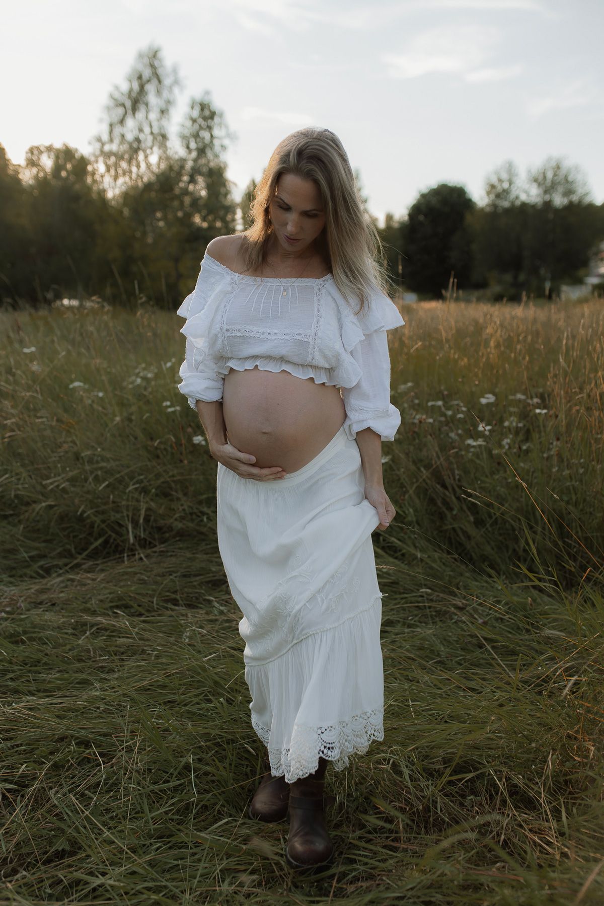 En gravid kvinna i spetsklänning som går på en sommaräng i solnedgången. Hon håller sin hand på magen och tittar ner på  magen.