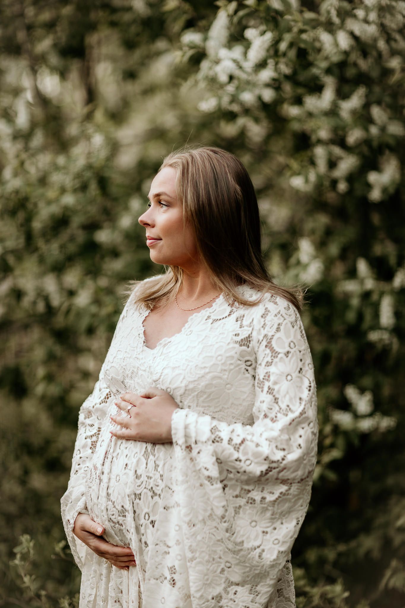 En gravid kvinna i bohemisk spetsklänning som står och tittar åt sidan. Bakom henne är ett grönt träd.