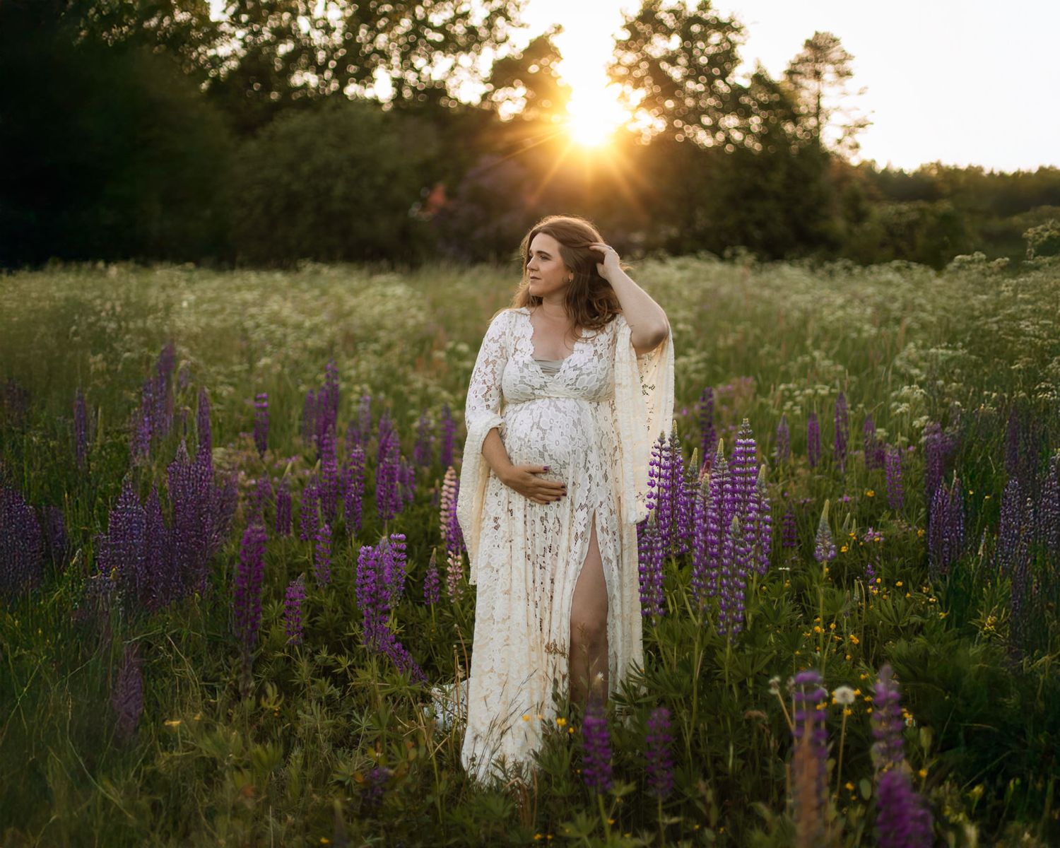 En gravid kvinna på en blommig sommaräng, klädd i bohemisk spetsklänning och hon håller sin hand i håret medan hon tittar åt sidan.