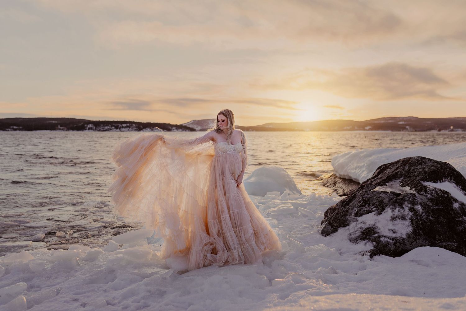 En gravid kvinna i en ljusrosa klänning står på isig klippa framför en havsvik, i solnedgången, med mjuka moln och berg i bakgrunden.