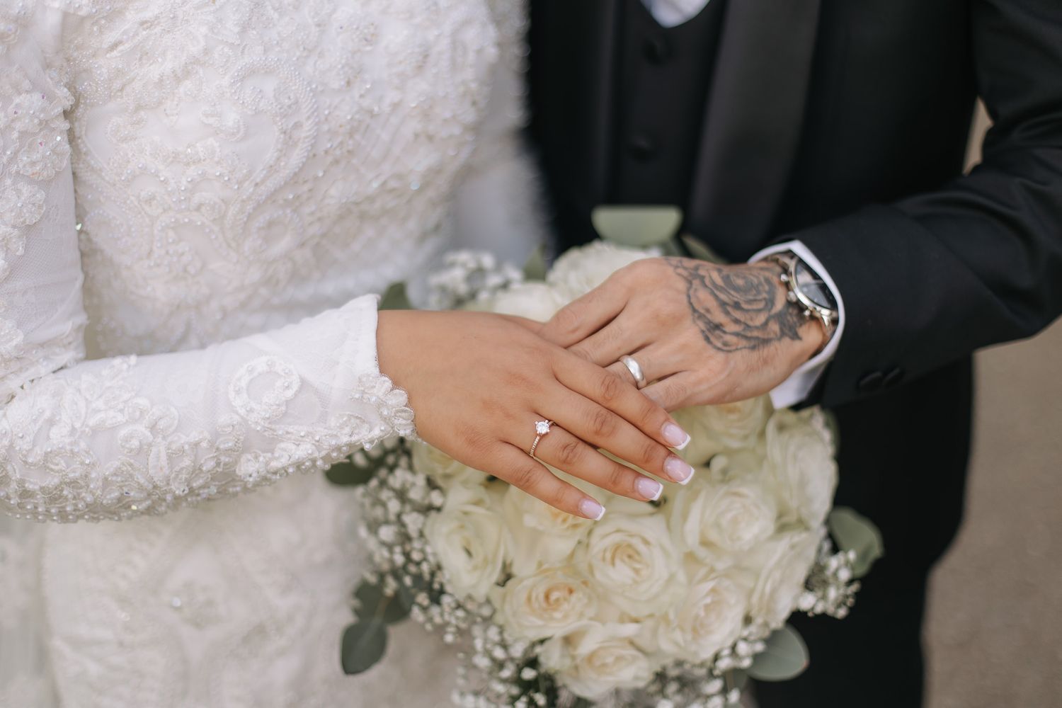 Närbild på ett brudpars händer, där ringarna är i fokus. Mannen har en tatuering på sin hand och de håller händerna ovanför brudbuketten.