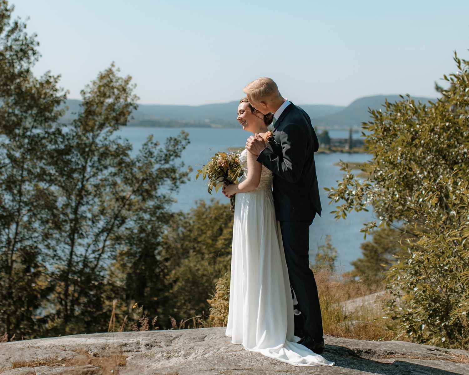En kostymklädd man och en kvinna i brudklänning som står på en klippa. Det är vatten och berg i bakgrunden. Kvinnan har en brudbukett.