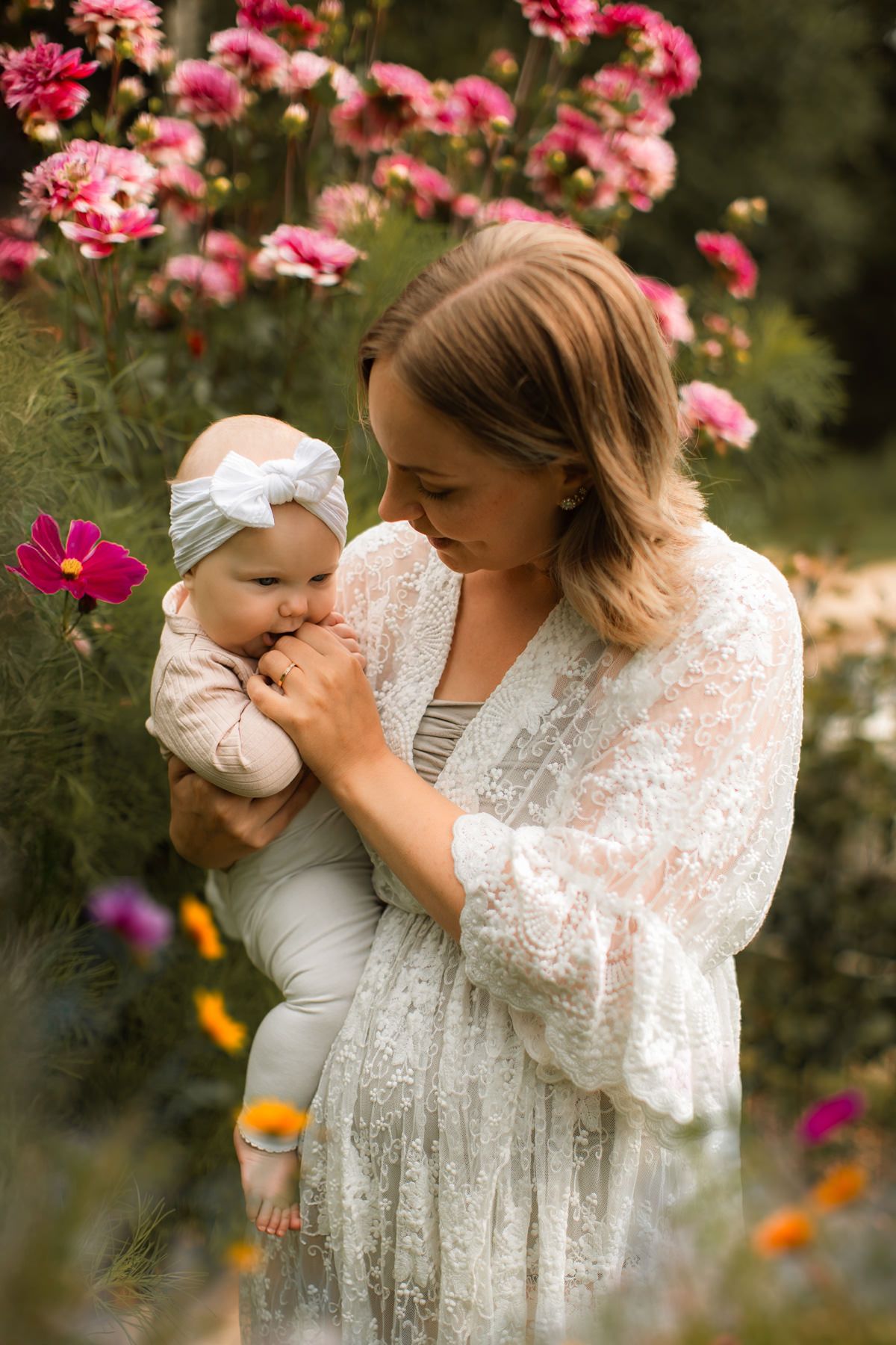 En kvinna klädd i bohemisk spetsklänning står framför en blommig buske. Hon håller en liten bebis i sin famn och hon ler mot bebisen.