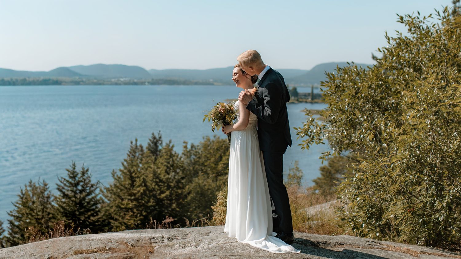 En man klädd i kostym och en kvinna klädd i brudklänning och en med brudbukett i sina händer. Mannen står bakom kvinnan och håller om henne.