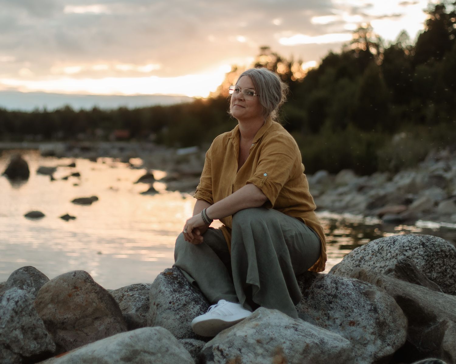 En kvinna som sitter på stenar med vatten, skog och en solnedgång i bakgrunden. Det är sommarkväll.