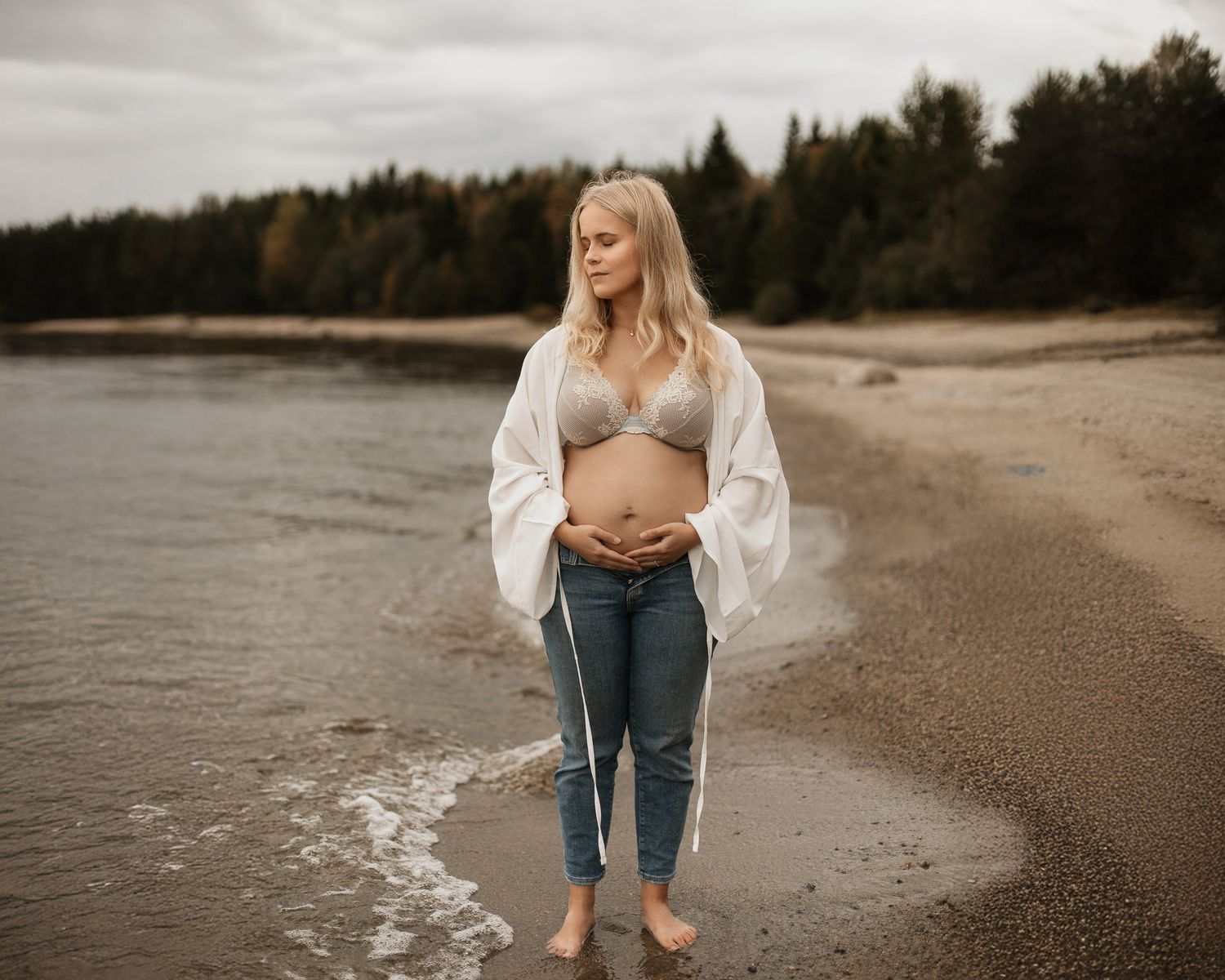 Gravid kvinna står i grunt vatten, iklädd bh och jeans, med en vit skjorta som är uppknäppt och en strand i bakgrunden.