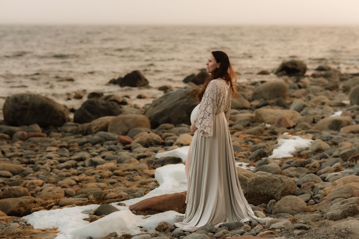 En gravid kvinna i en vintageklänning, står på en stenig strand, med utsikt över havet, i solnedgången, med ett mjukt gyllene ljus.