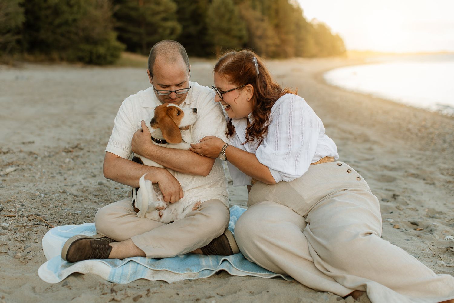 En man och en kvinna klädda i ljusa linnekläder sitter på en sandstrand i kvällssolen. Mannen håller i en hund som tittar på kvinna.
