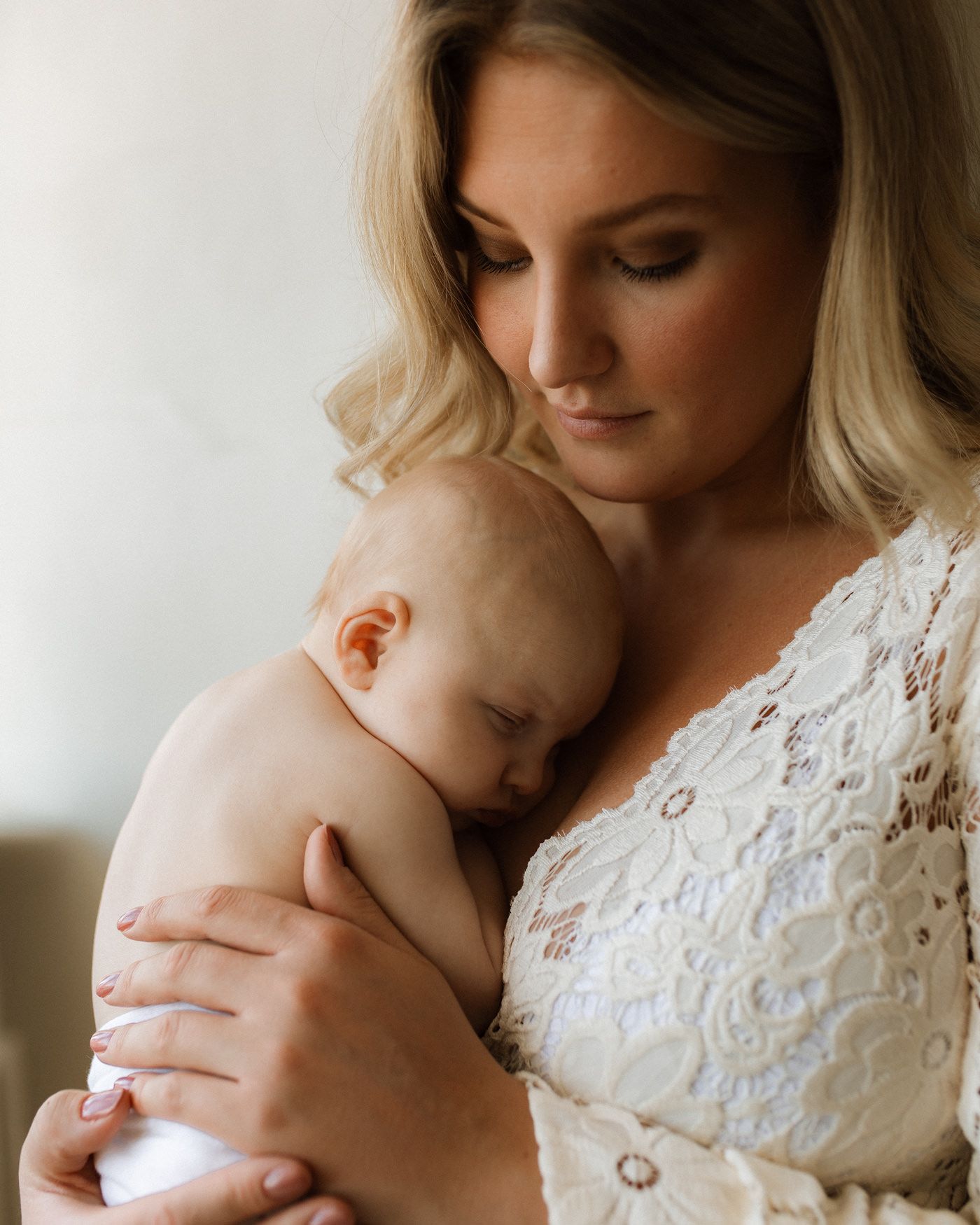 En kvinna med bohemisk spetsklänning som håller i en liten nyfödd bebis i sin famn. Bebisen sover mot mammans bröst och mamman ser på babyn.