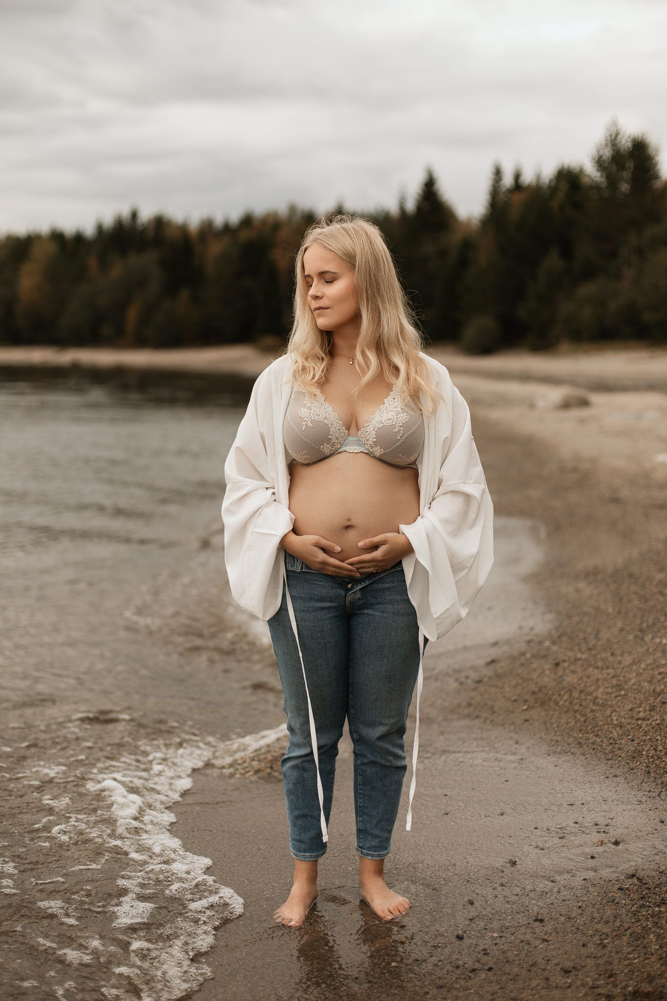 En gravid kvinna står barfota vid vattenbrynet, iklädd en spets-bh och jeans, med en vit öppen skjorta, mot en molnig bakgrund.
