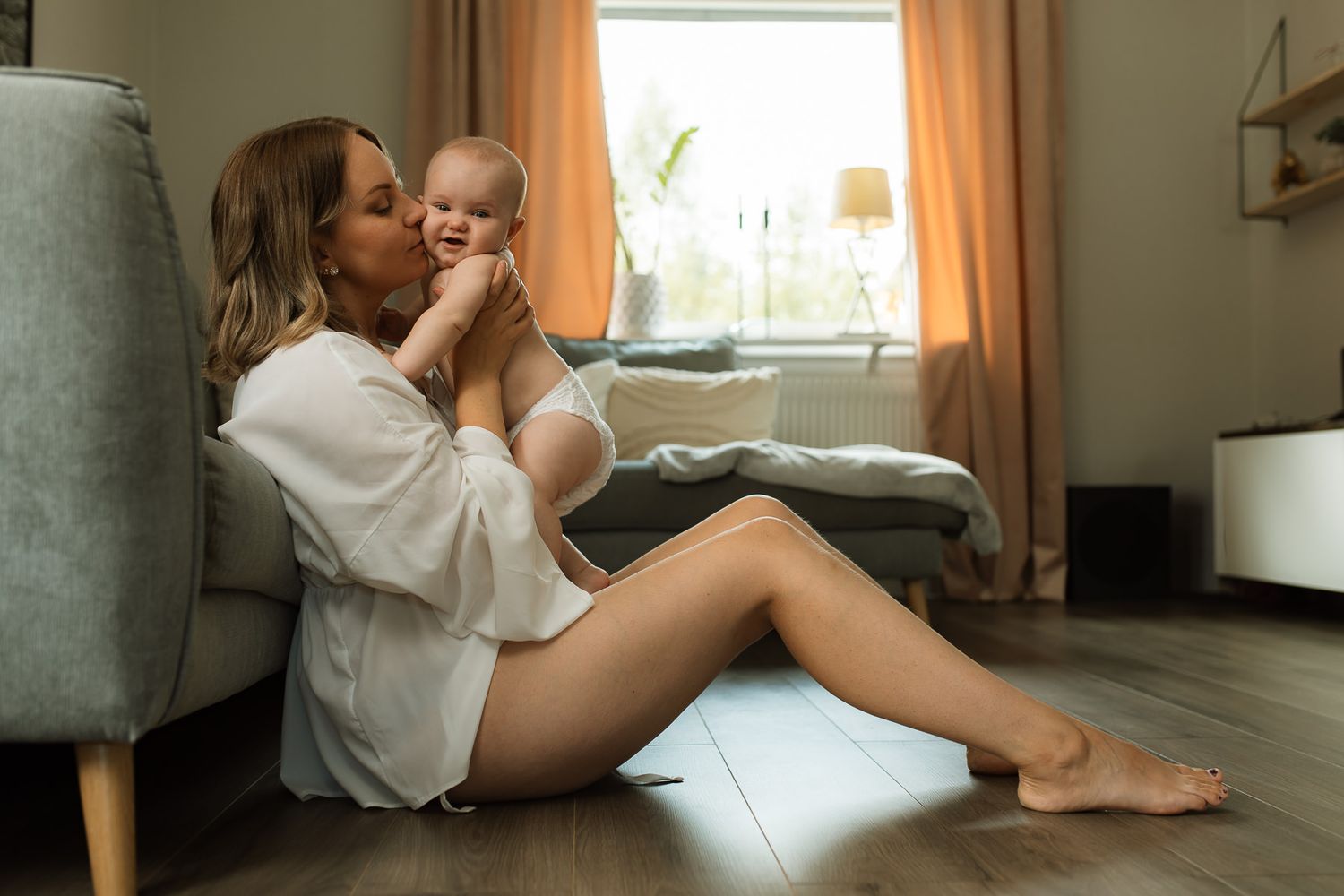 En kvinna som sitter på golvet med bara ben i en vit stor skjorta och som håller i en bebis i sin famn. Hon pussar bebisen på kinden.