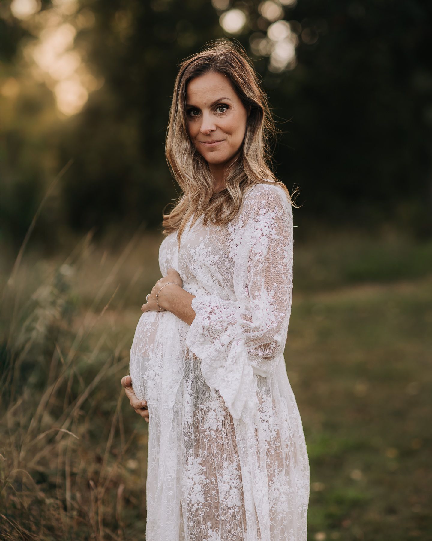 En gravid kvinna klädd i skir spetsklänning står i ett grönt område med en solnedgång i bakgrunden. Hon håller sina händer kring sin mage.