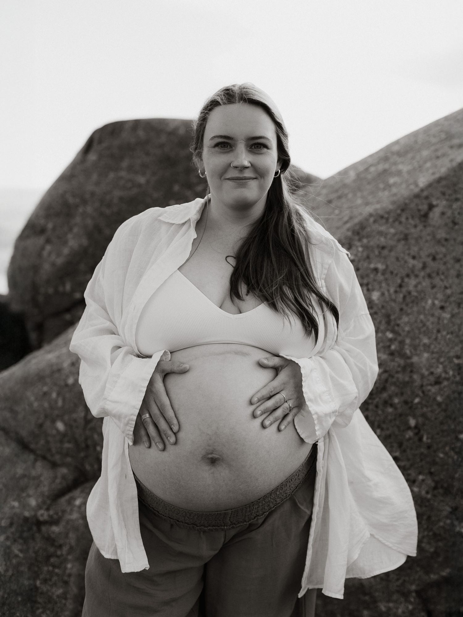 En gravid kvinna med öppen skjorta och synlig bh står på en klippa. Hon håller sina händer på magen och tittar in i kameran.