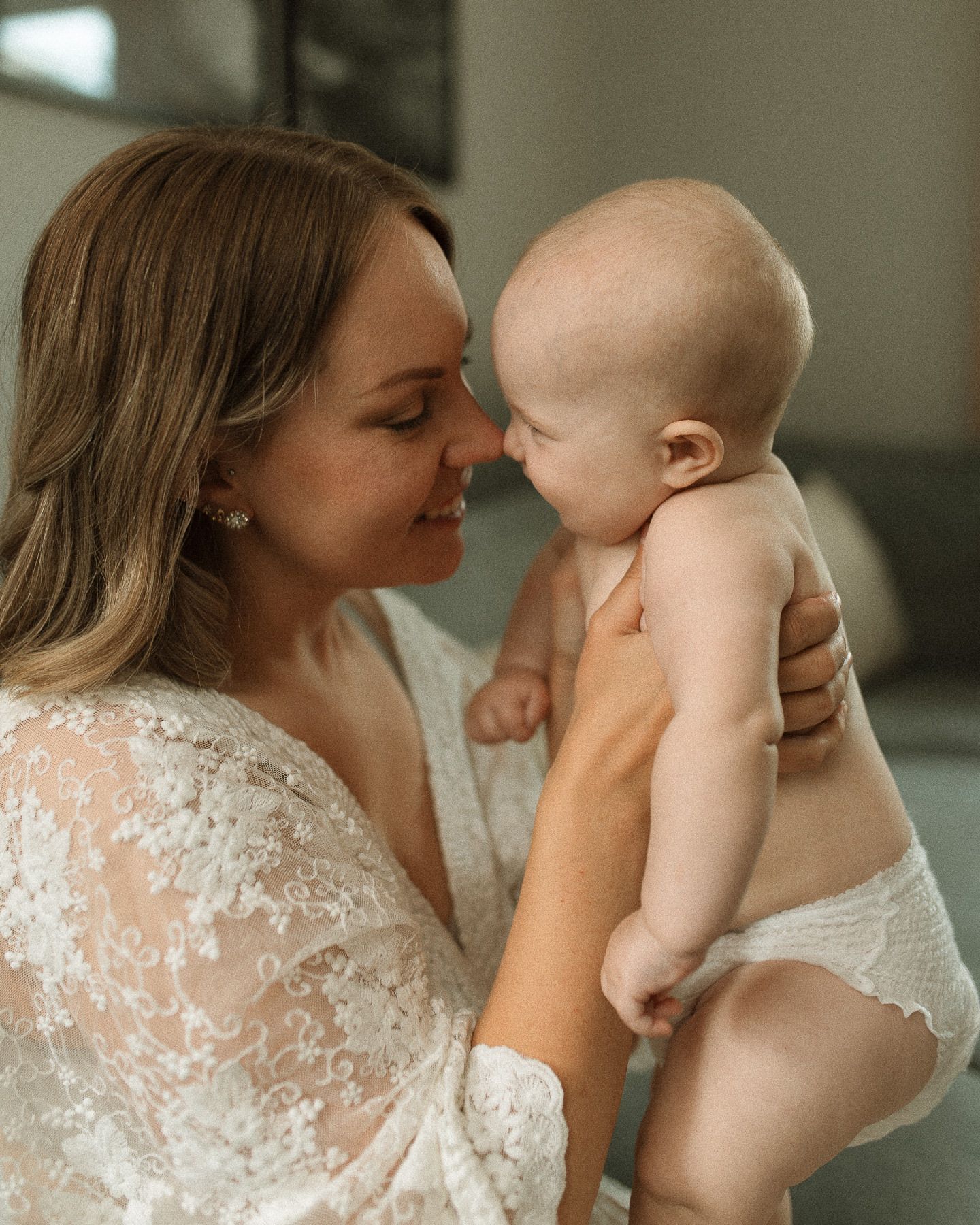 En mamma i en vit spetstopp som håller en bebis med blöja i en mysig inomhusmiljö. De håller näsorna mot varandra.