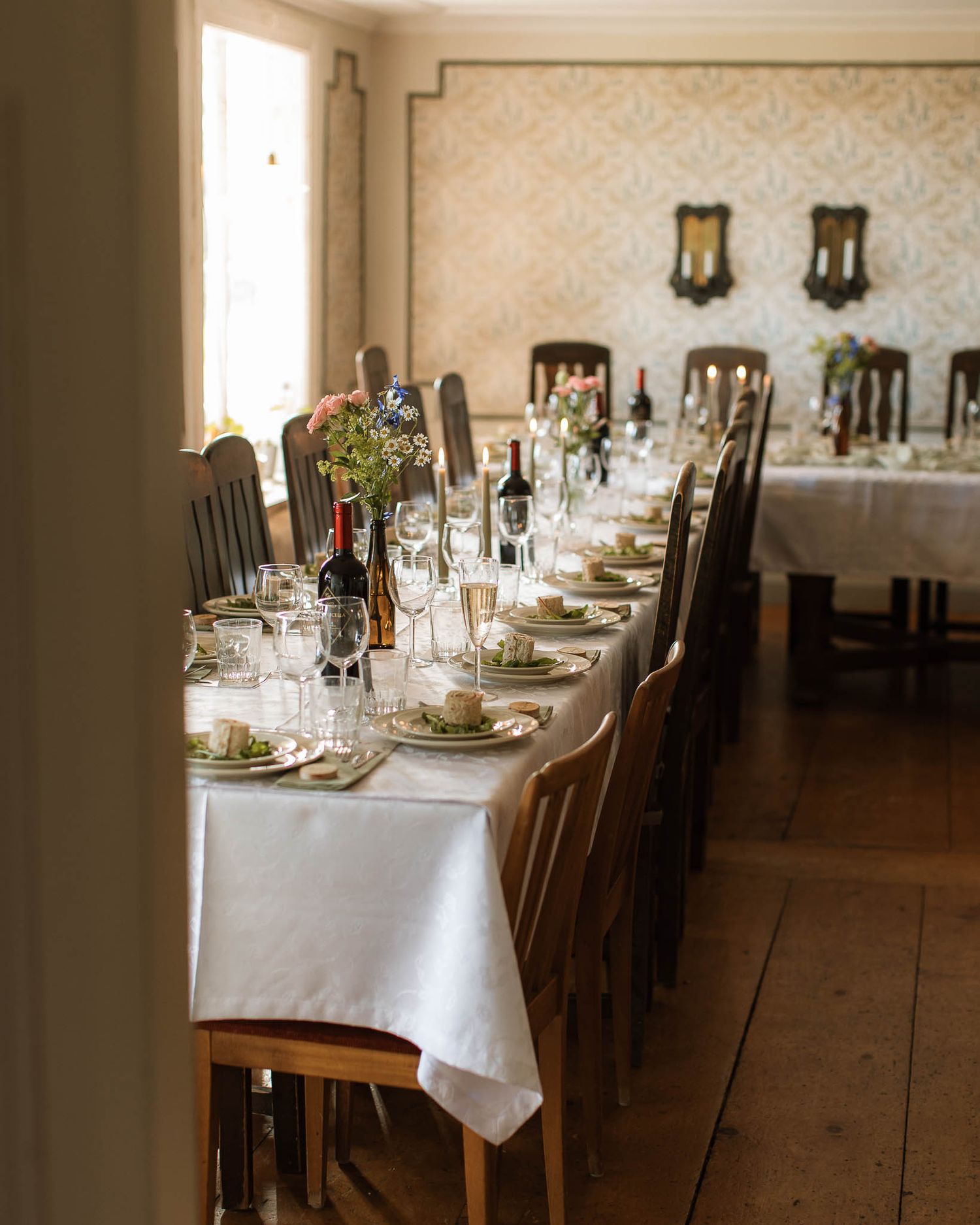 Ett gammalt rum med ett vackert dukat bord. Det står tallrikar, glas, servetter, blommor och vinflaskor på bordet.