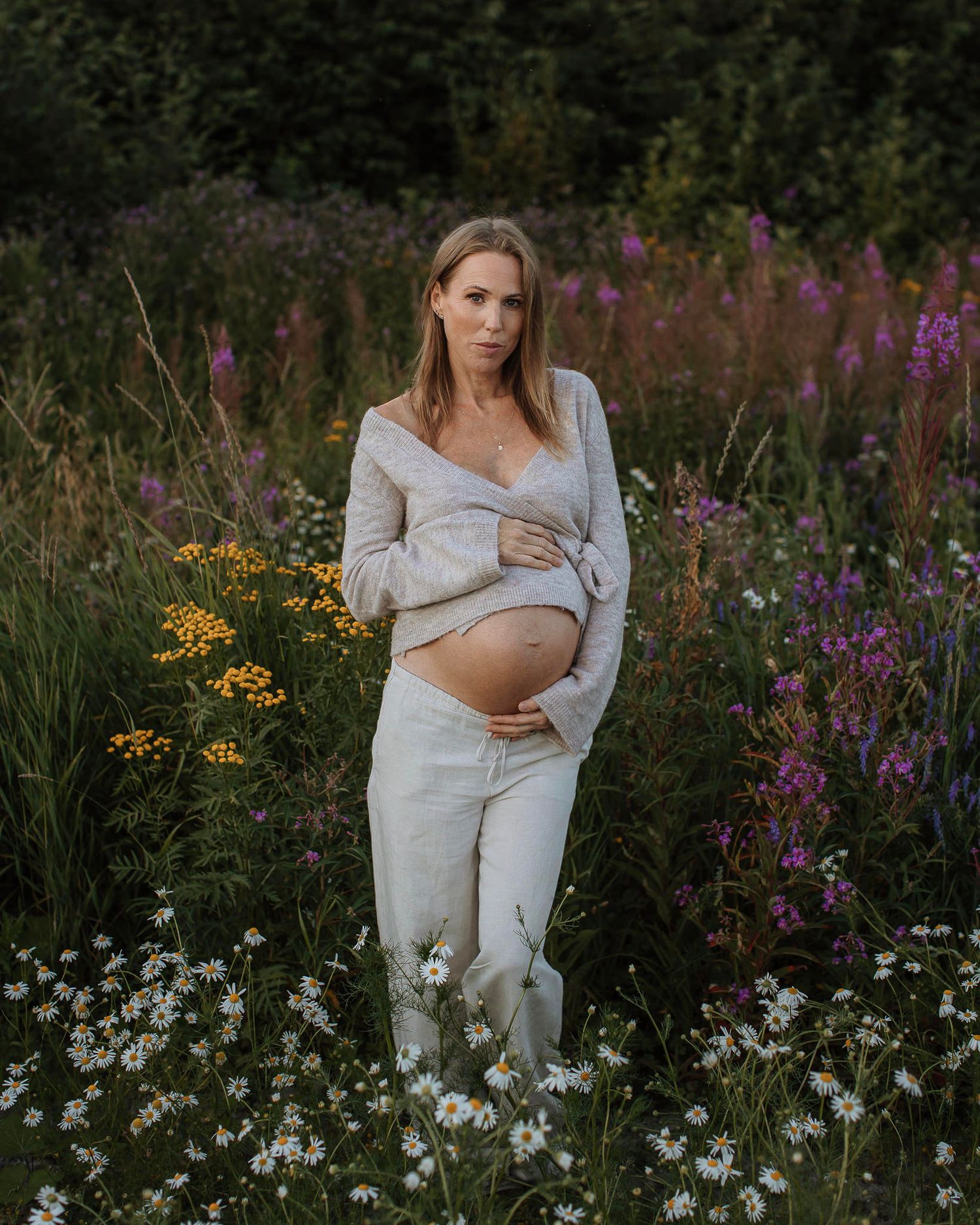En gravid kvinna med långt hår som står på en blomsteräng med färgglada blommor. Hon håller sina händer om sin mage.