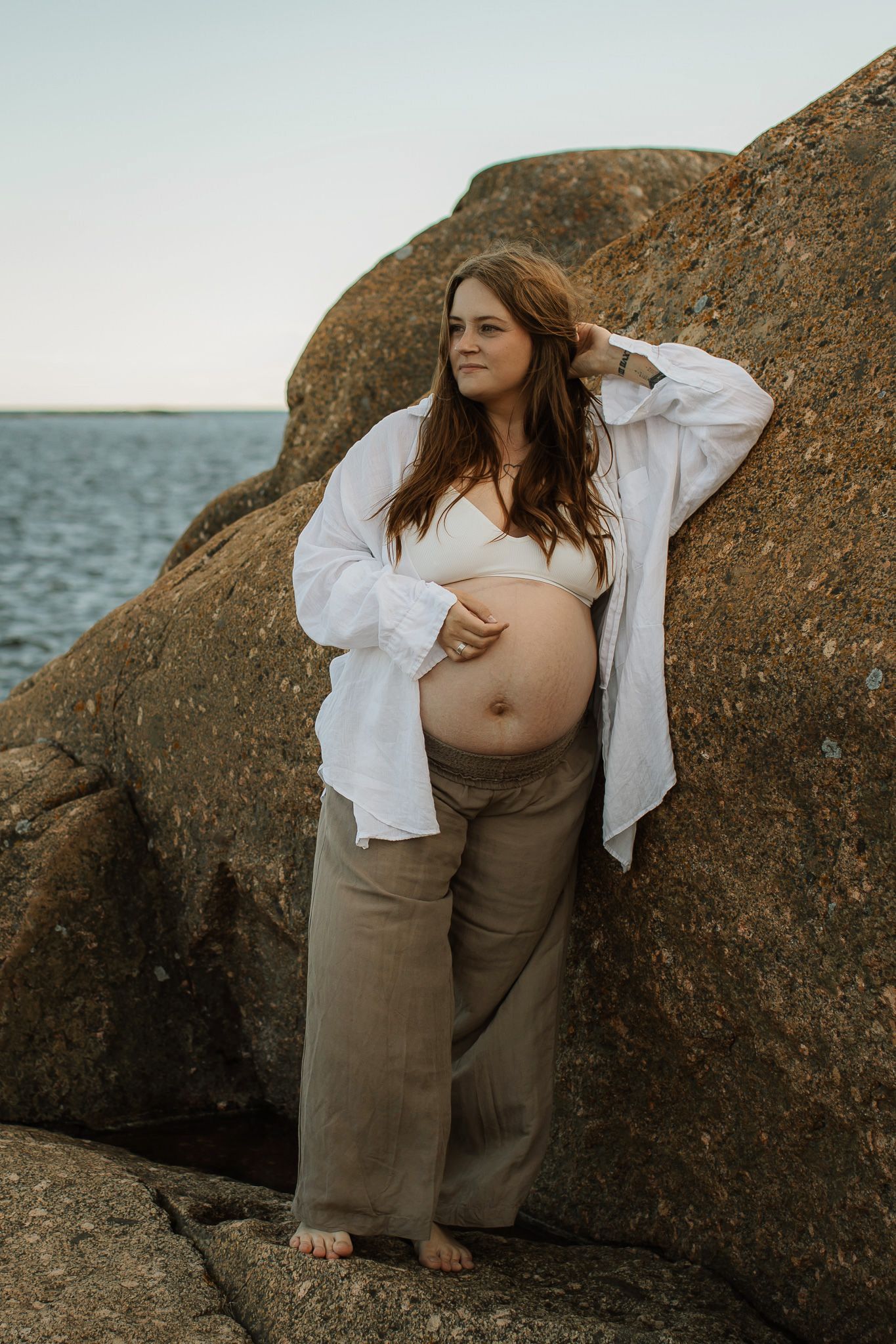 En gravid kvinna i vit skjortat och linnebyxor som står lutad mot en klippa. Hon håller ena handen på sin mage.