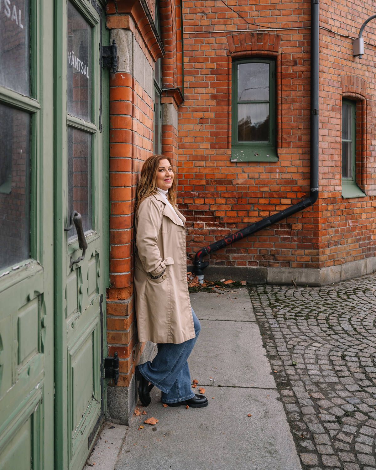 En kvinna i beige trenchcoat, jeans och långt hår. Hon håller händerna i fickorna och lutar sig mot en tegelvägg.